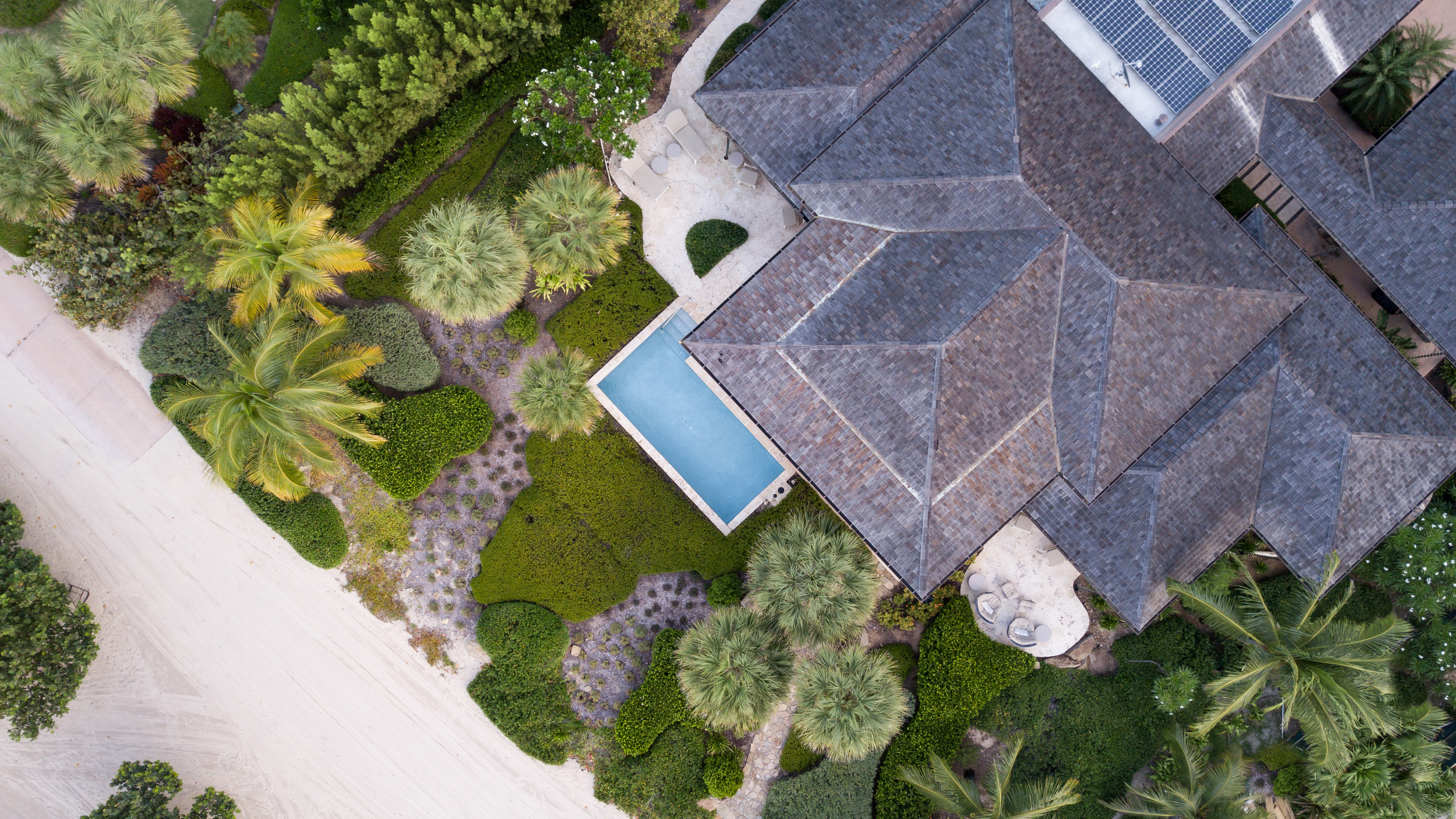 Aerial view of Cheemaun Villa with its intricate roof design, a small backyard pool, and lush landscaping featuring various trees and shrubs. The property is bordered by a sandy path on one side.