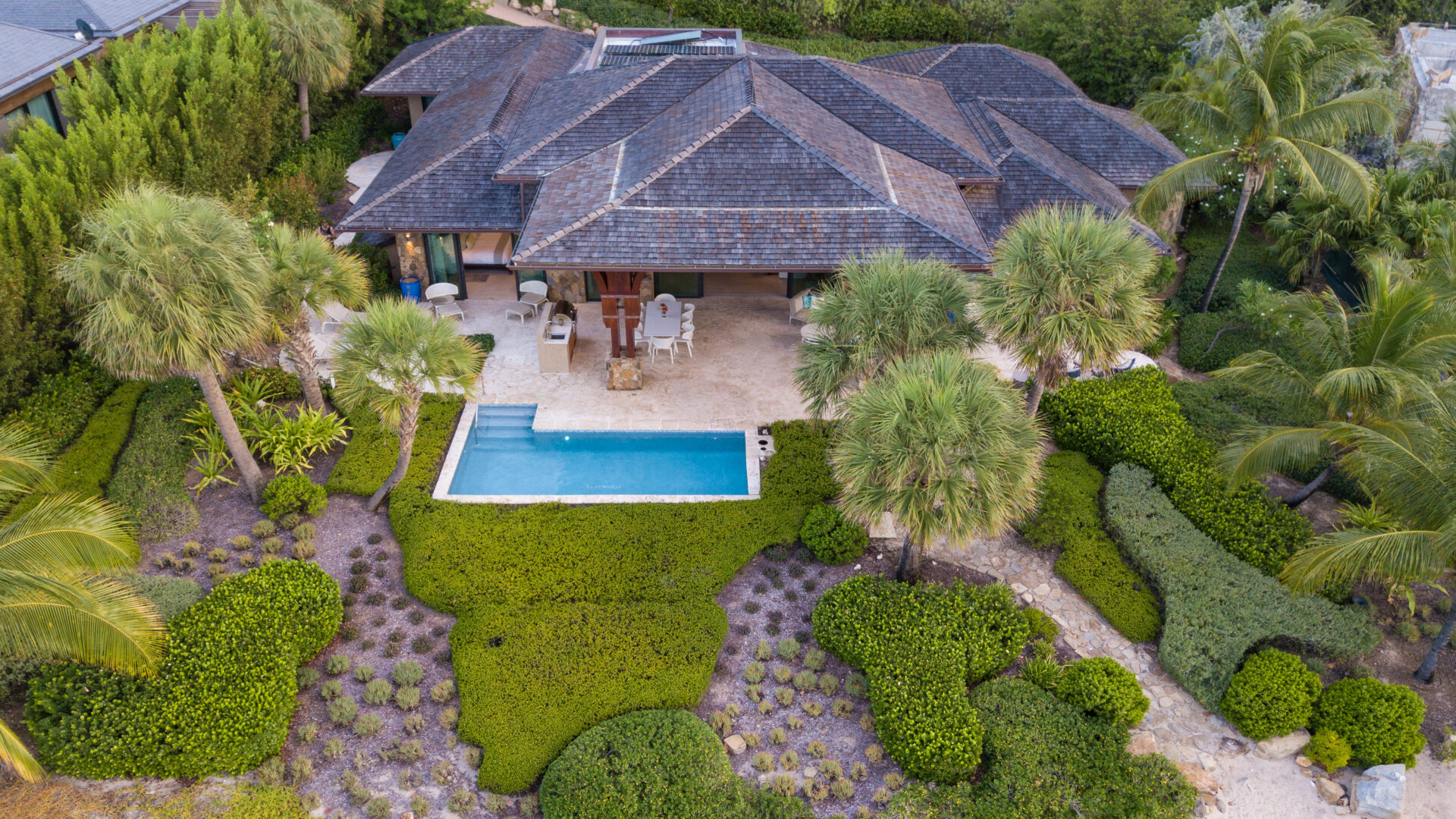 Aerial view of the luxurious Cheemaun Villa surrounded by lush greenery. The villa features a thatched roof and an outdoor patio with lounge chairs, leading to a rectangular swimming pool. Palm trees and manicured bushes enhance the serene, verdant setting.