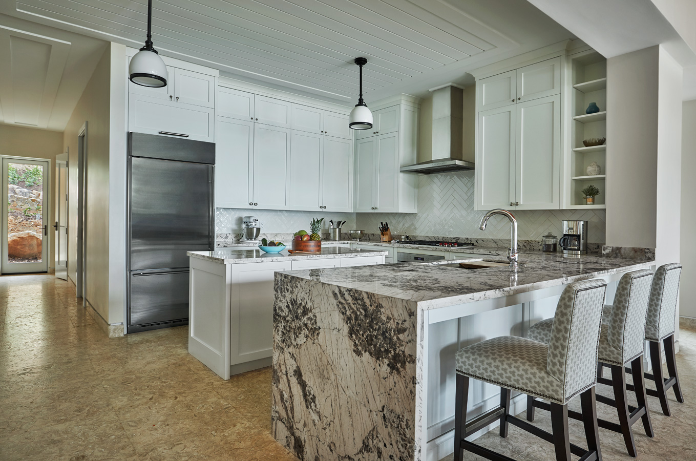 A contemporary kitchen in Cheemaun Villa with white cabinets, a stainless steel refrigerator, and a marble countertop island. The island features a gray and white marble pattern and is accompanied by two light-patterned upholstered barstools. Pendant lights hang overhead.