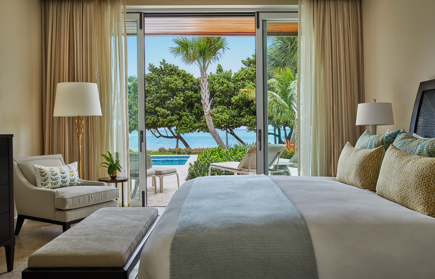 A luxurious villa bedroom features a neatly made bed with patterned pillows and a light blue throw. Large sliding glass doors reveal a private patio with chairs, tropical greenery, and a glimpse of the serene Cheemaun blue ocean. Soft lighting and neutral tones create a calm ambiance.