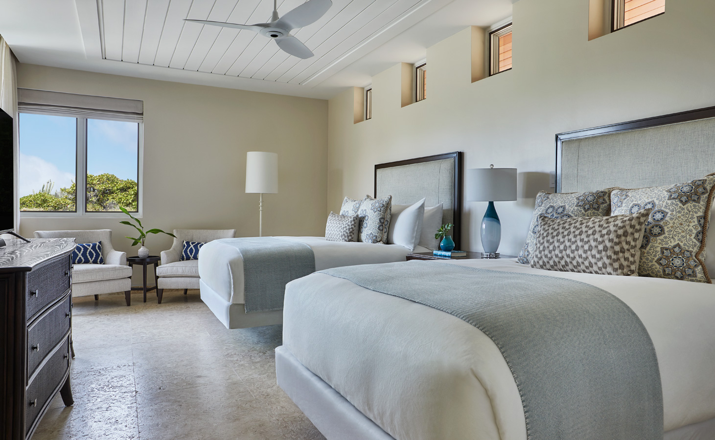 A modern villa bedroom with two neatly made beds featuring gray and white bedding, a dark wooden dresser, and a sitting area with a white chair. A window with a view of greenery is on the left, complemented by several small Cheemaun windows near the ceiling and a ceiling fan above.