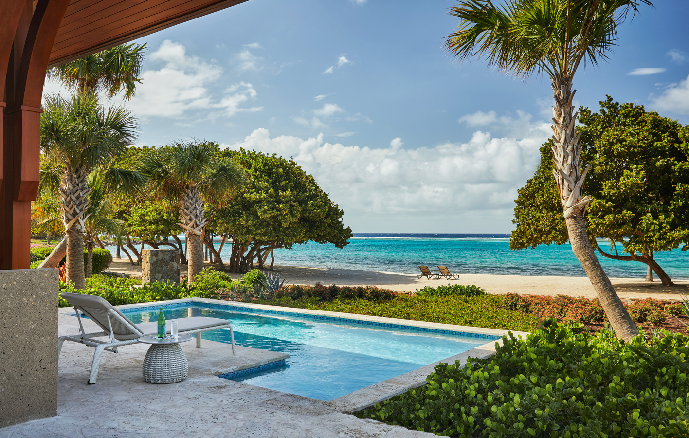 A serene beach scene at Cheemaun Villa with a private pool in the foreground, flanked by poolside loungers and a small table. The backdrop boasts palm trees, lush greenery, and a sandy beach leading to a turquoise ocean under a partly cloudy sky. Two sun loungers rest on the shore facing the sea.