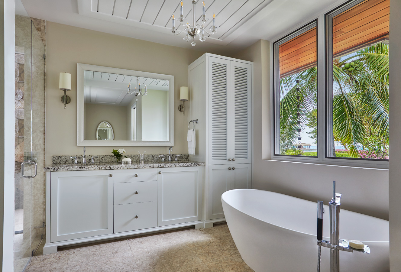 A modern bathroom in Villa Cheemaun with light gray walls, featuring a large mirror above a double-sink white vanity with a granite countertop, a freestanding white bathtub, and a window with a view of palm trees. A chandelier hangs from the ceiling, and the floor is tiled.