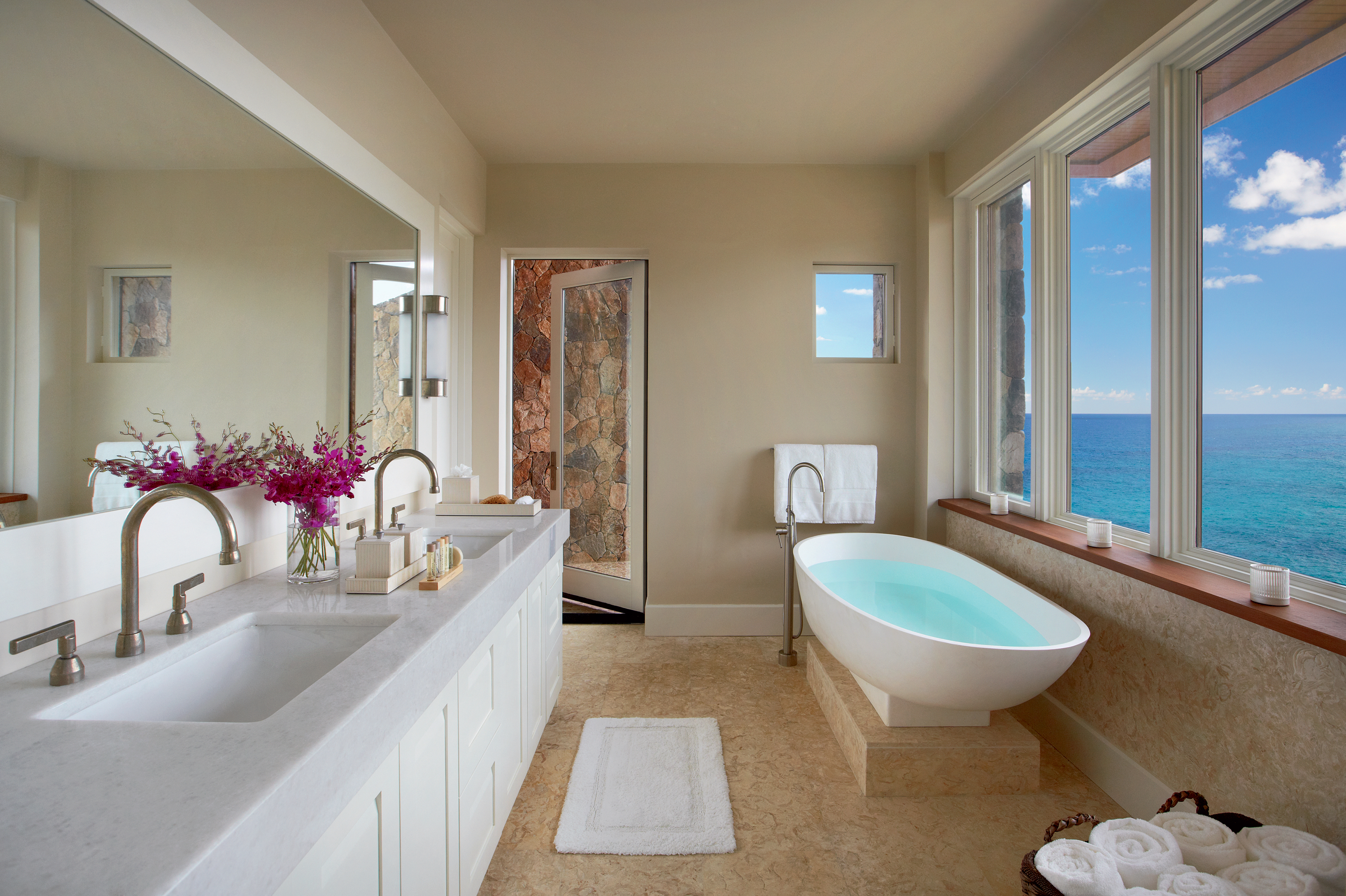 Spacious bathroom at The Cliff Suites with a large window giving an ocean view. The room includes a double sink vanity with flowers, a freestanding white bathtub next to the window, folded white towels, and neutral-toned decor with a small, partly open door revealing a stone wall.