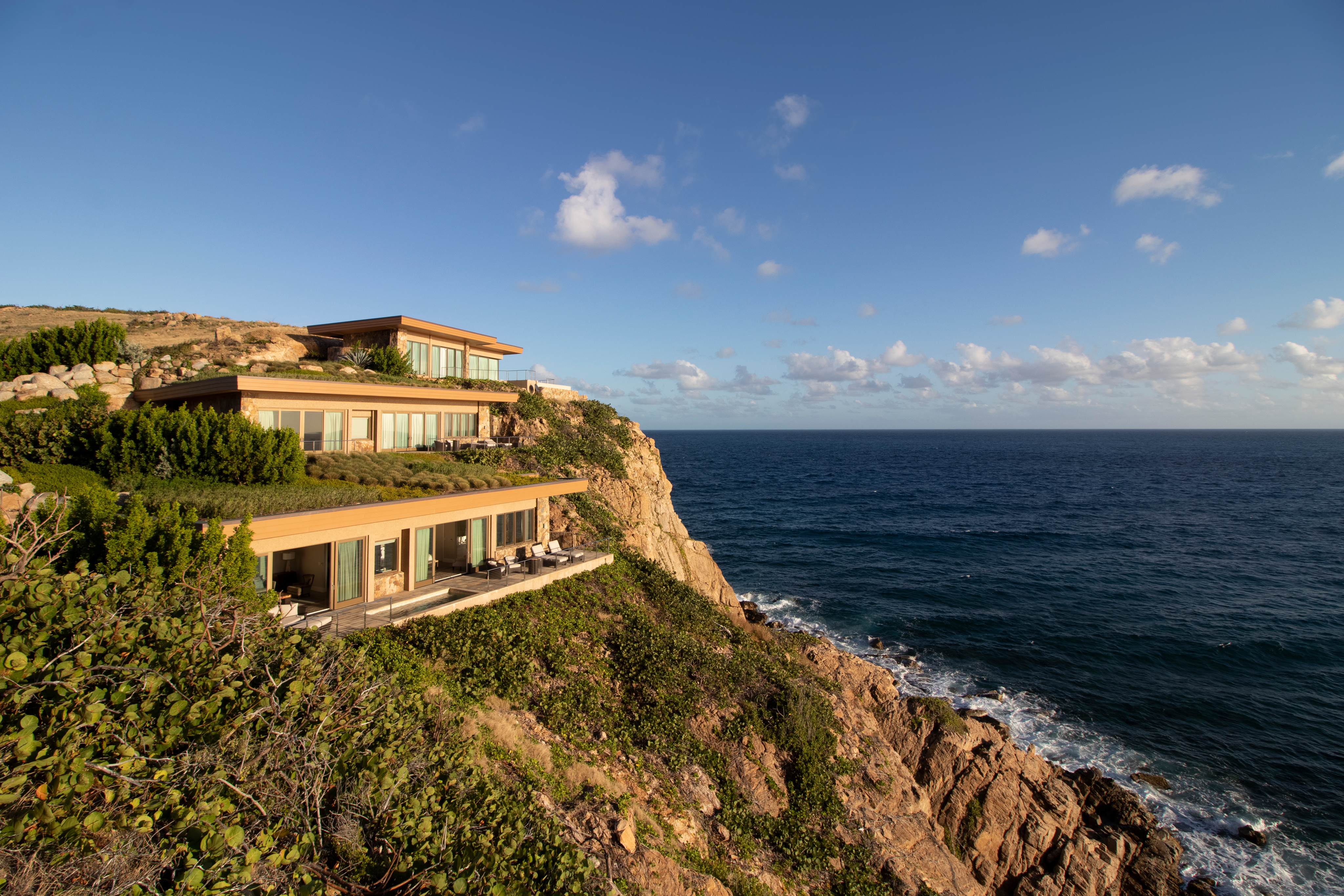 The Cliff Suites, a modern, multi-story coastal house with large windows and terraces, is perched on a rocky cliffside overlooking the ocean. The sun casts a warm light on the house and surrounding vegetation. The sky is clear with a few scattered clouds.