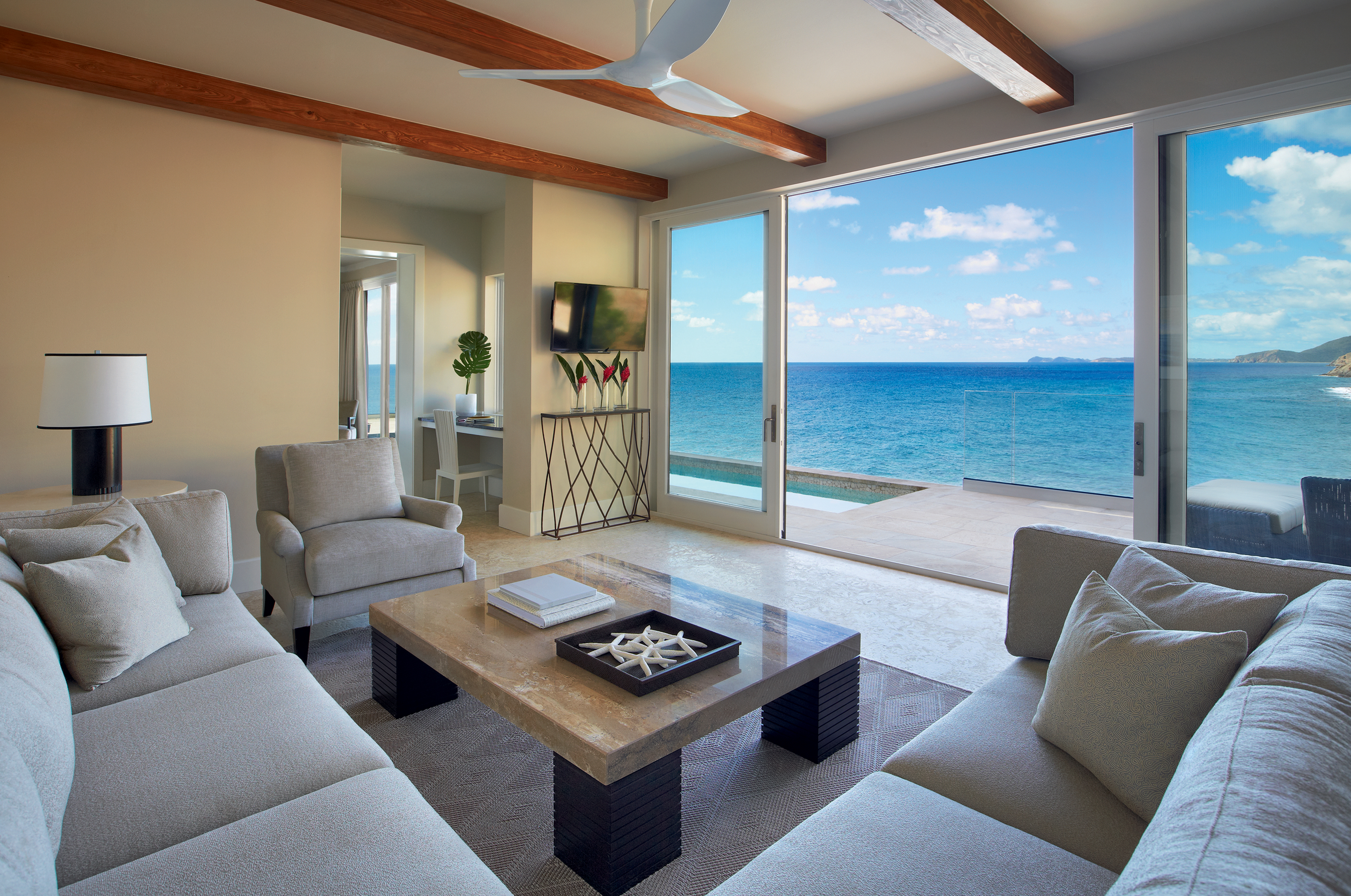 Spacious living room at The Cliff Suites with a large beige sectional sofa, two armchairs, and a glass-top coffee table. The room features ceiling beams, a mounted TV, and large sliding glass doors that open to an ocean view with blue skies and scattered clouds.