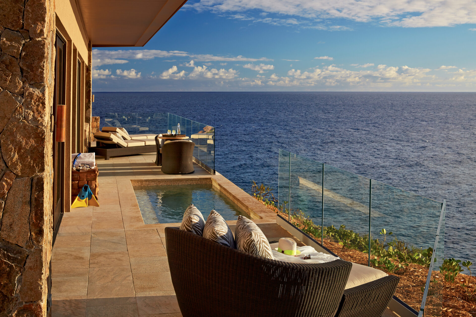 The Cliff Suites' luxurious seaside terrace features a cozy seating area with striped pillows, a small plunge pool, and glass railings offering an unobstructed ocean view under a partly cloudy sky. The scene is tranquil and inviting, perfect for those seeking premium luxury accommodation.