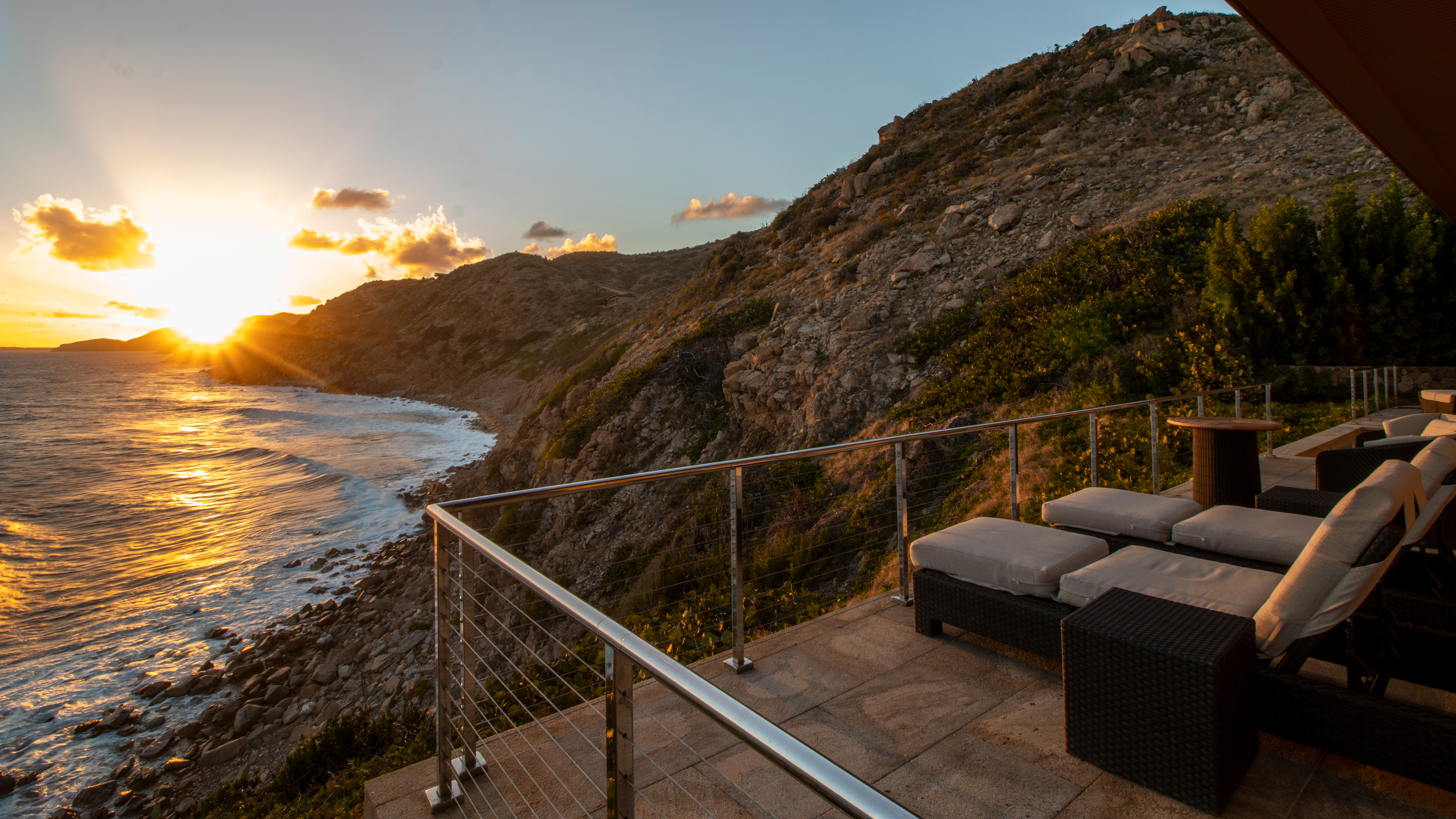 A scenic coastal view at sunset with rocky cliffs meeting the ocean. The foreground features an outdoor lounge area with cushioned seating and a glass railing at The Cliff Suites. The horizon glows with the setting sun casting a warm light over the landscape.