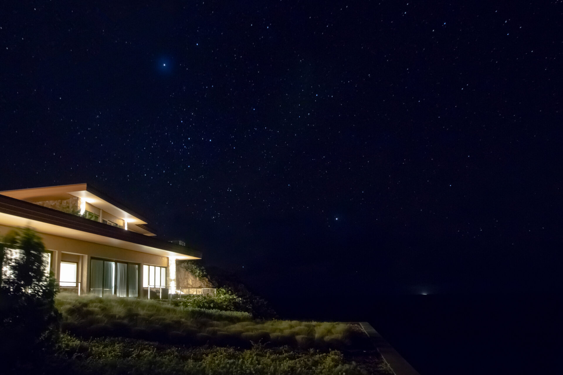 A modern house with large windows, part of The Cliff Suites, is illuminated at night under a starry sky. The vast expanse of stars twinkles above, enhancing the serene and peaceful atmosphere. The house is surrounded by shrubbery, and there is a faint light in the distant horizon.