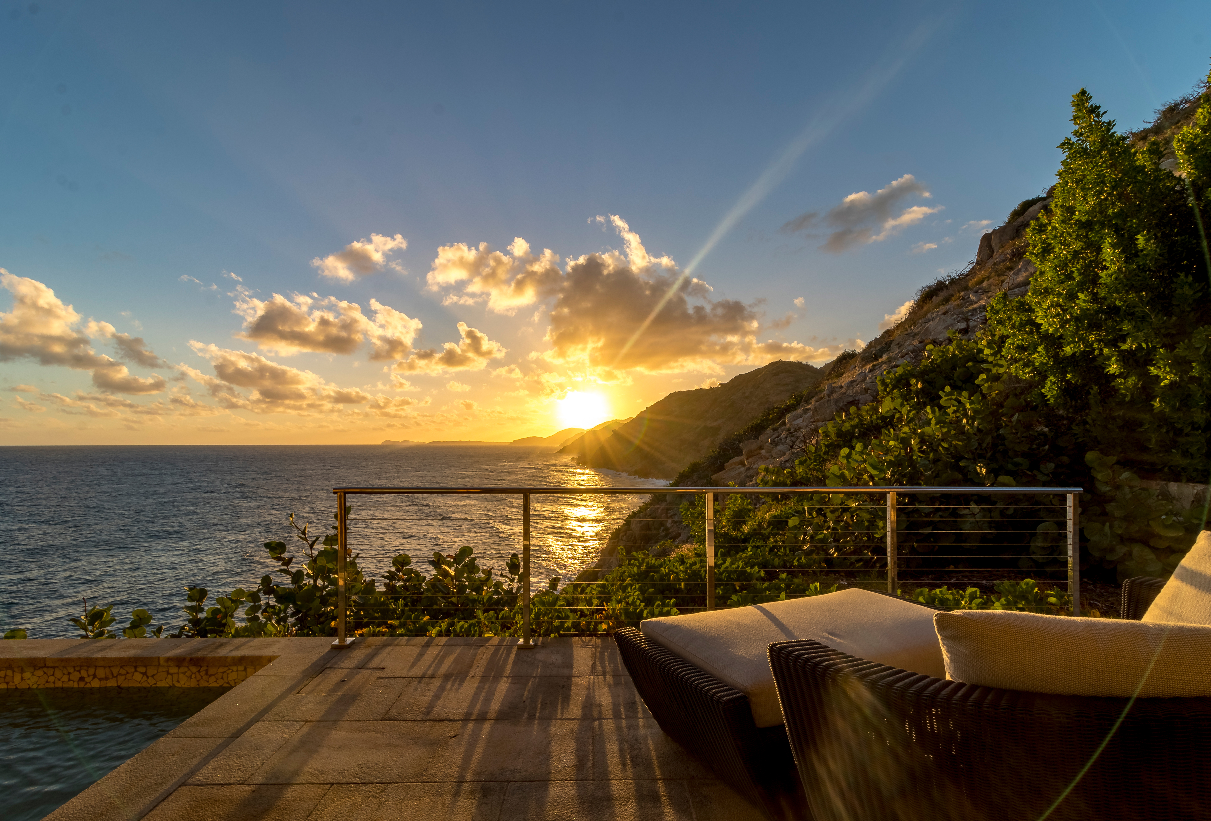 A scenic view of a beach at sunset, featuring a calm ocean, a rugged cliff, and a sunlit sky with scattered clouds. At The Cliff Suites, relax in a wicker lounge chair with a white cushion on the patio in the foreground, bordered by lush plants and an elegant metal railing.