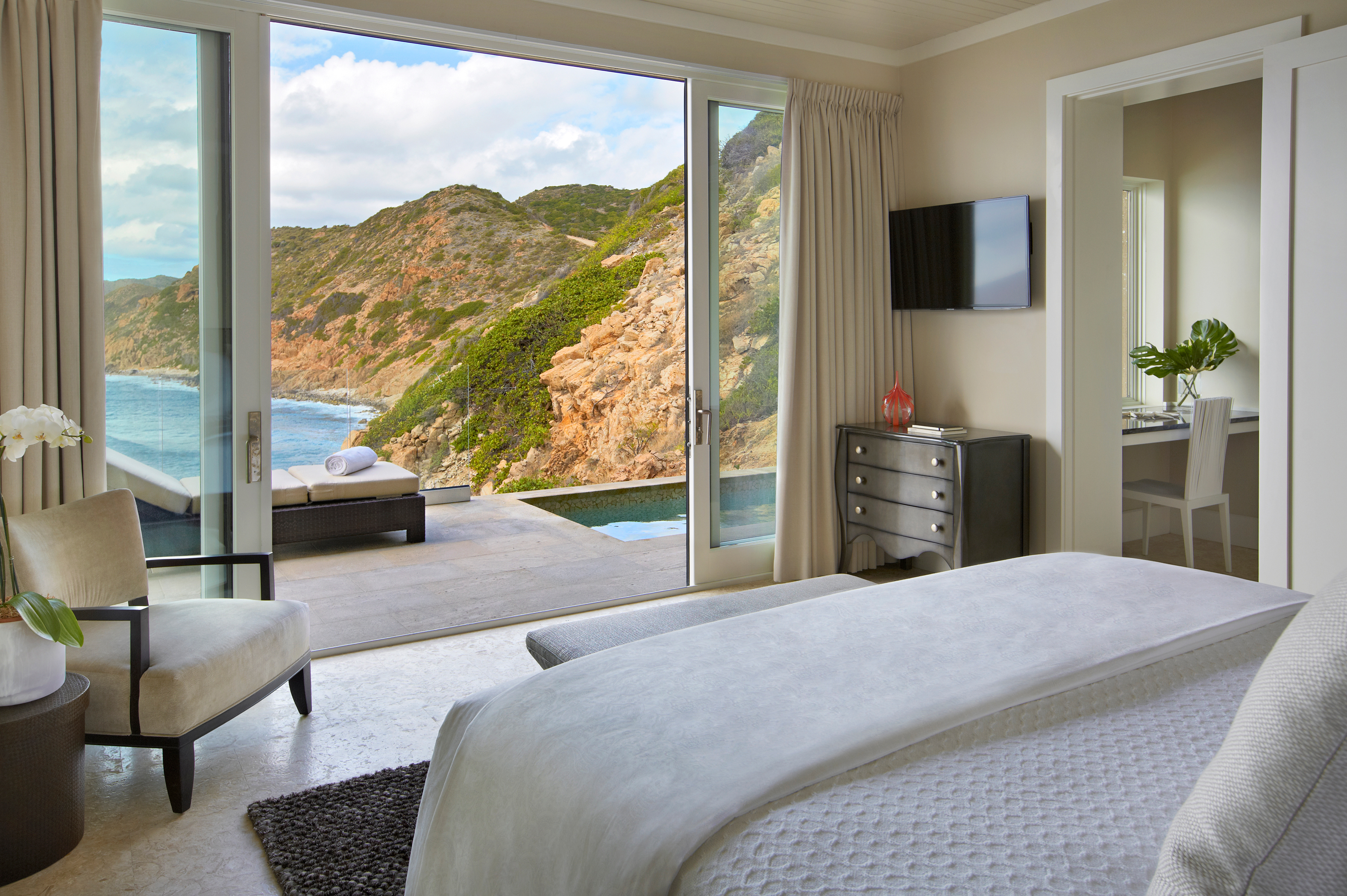 A luxurious bedroom at The Cliff Suites boasts glass sliding doors that open to a patio with lounge chairs, offering a scenic view of rocky cliffs and the ocean. The room features a neatly made bed, a wall-mounted TV, a chair near the window, and a dresser with a plant on top.