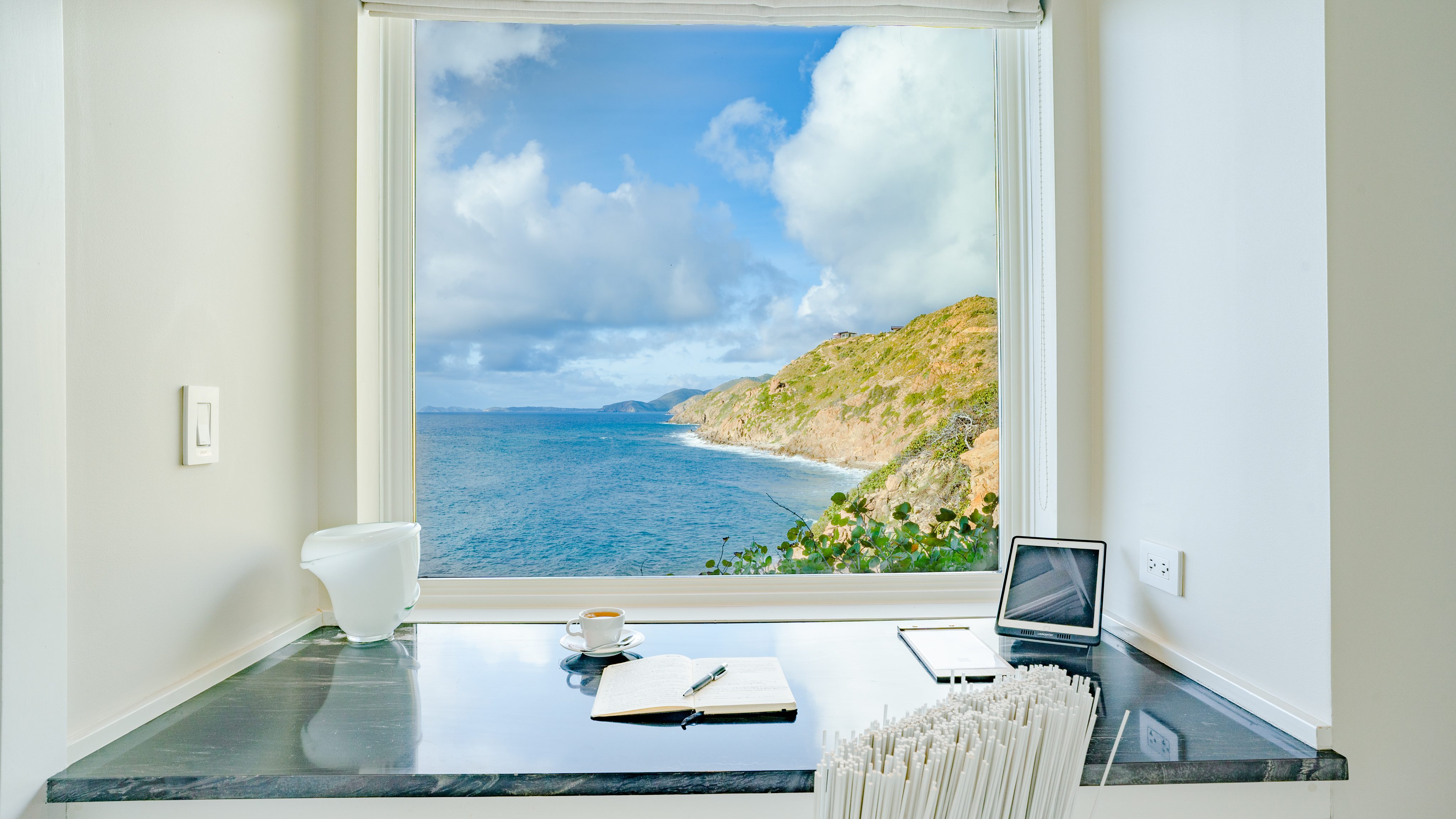 A serene workspace at The Cliff Suites offers a view of the ocean and rugged coastline through a large window. The desk features a laptop, a cup of coffee, an open notebook with a pen, a small light, and a power outlet. Bright, natural light fills the room.