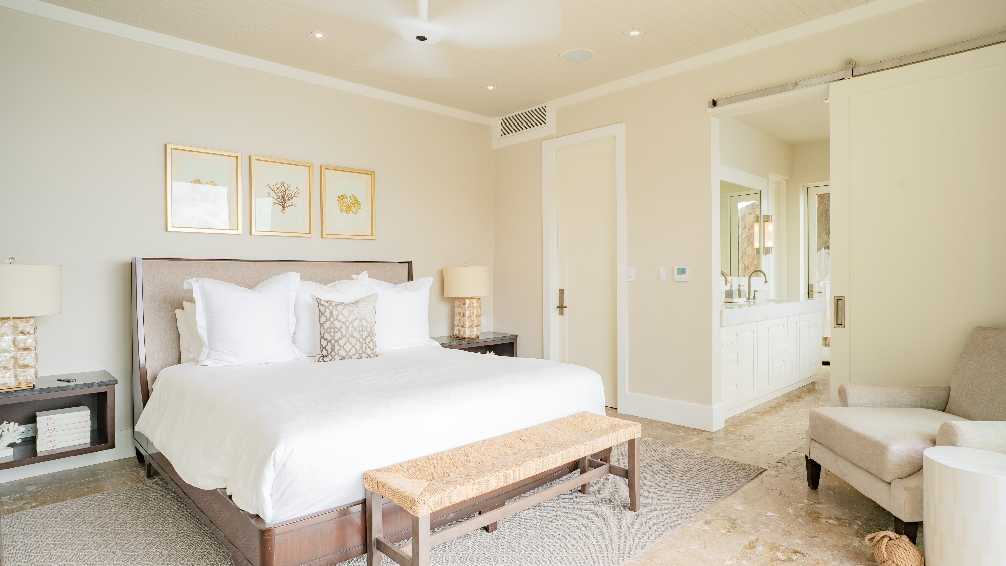 A bright and airy bedroom at The Cliff Suites features a large bed with white linens and decorative pillows. Two bedside tables with lamps flank the bed. Above the headboard are three framed art pieces. An armchair sits near the sliding door, revealing a glimpse of a bathroom.