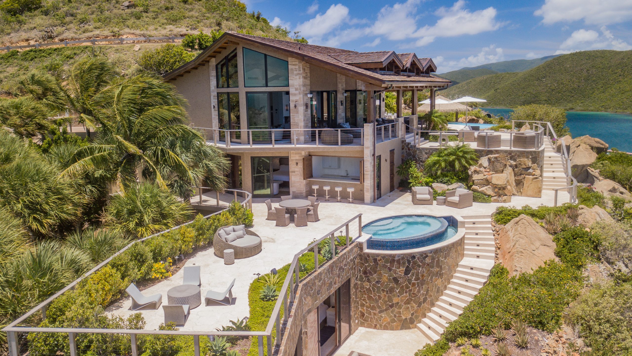 Compass Rose Villa is a luxurious, modern two-story residence overlooking a scenic body of water, nestled amidst lush greenery. The villa features large glass windows, multiple outdoor seating areas, a circular pool, and a spacious terrace with panoramic views of the surrounding hills and sea.