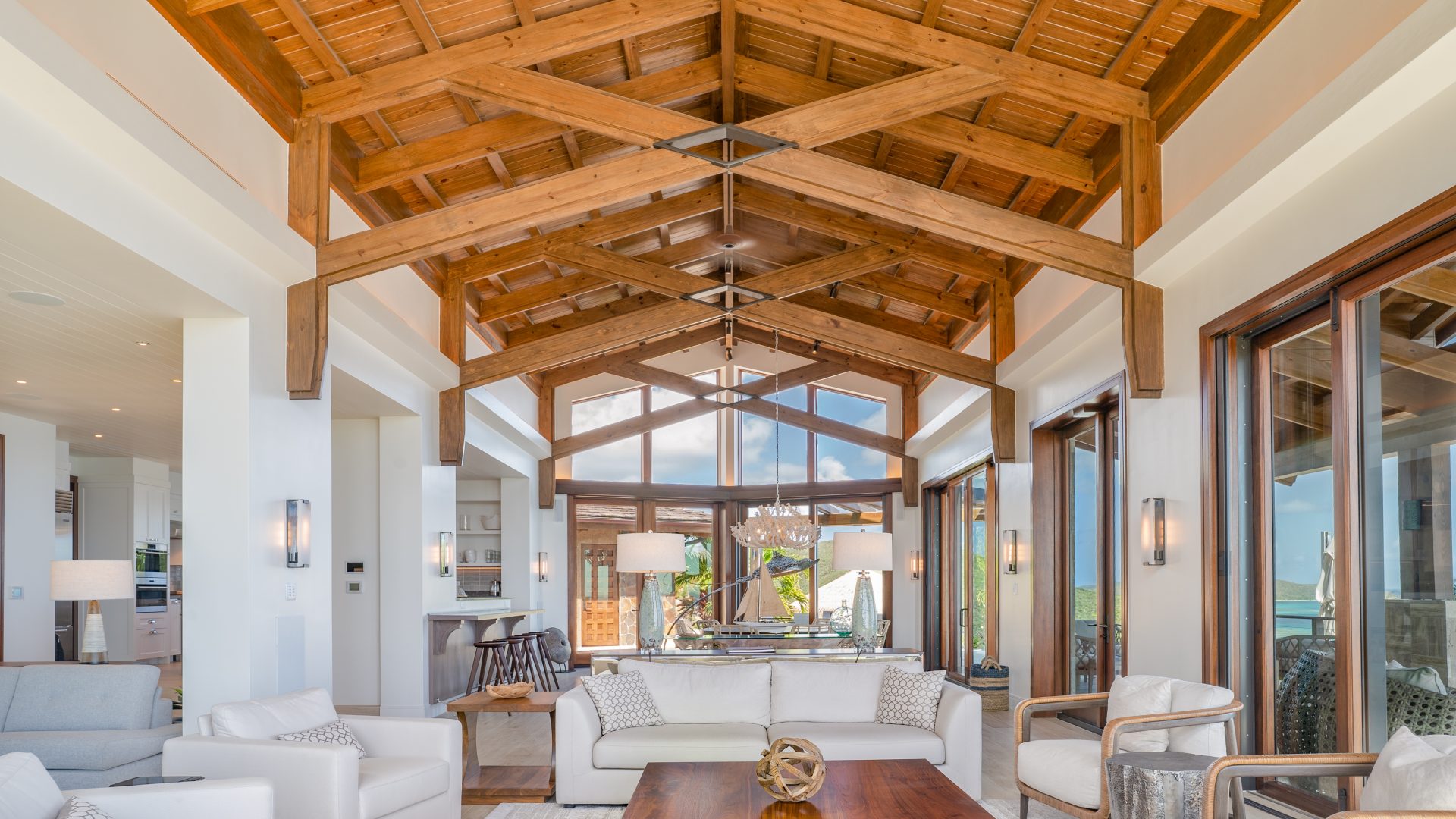 Compass Rose Villa boasts a spacious, modern living room with high, wooden-beamed ceilings. The room features large windows allowing ample natural light, white sofas, a wooden coffee table, and contemporary decor. Sliding glass doors lead to an outdoor space with a scenic view.