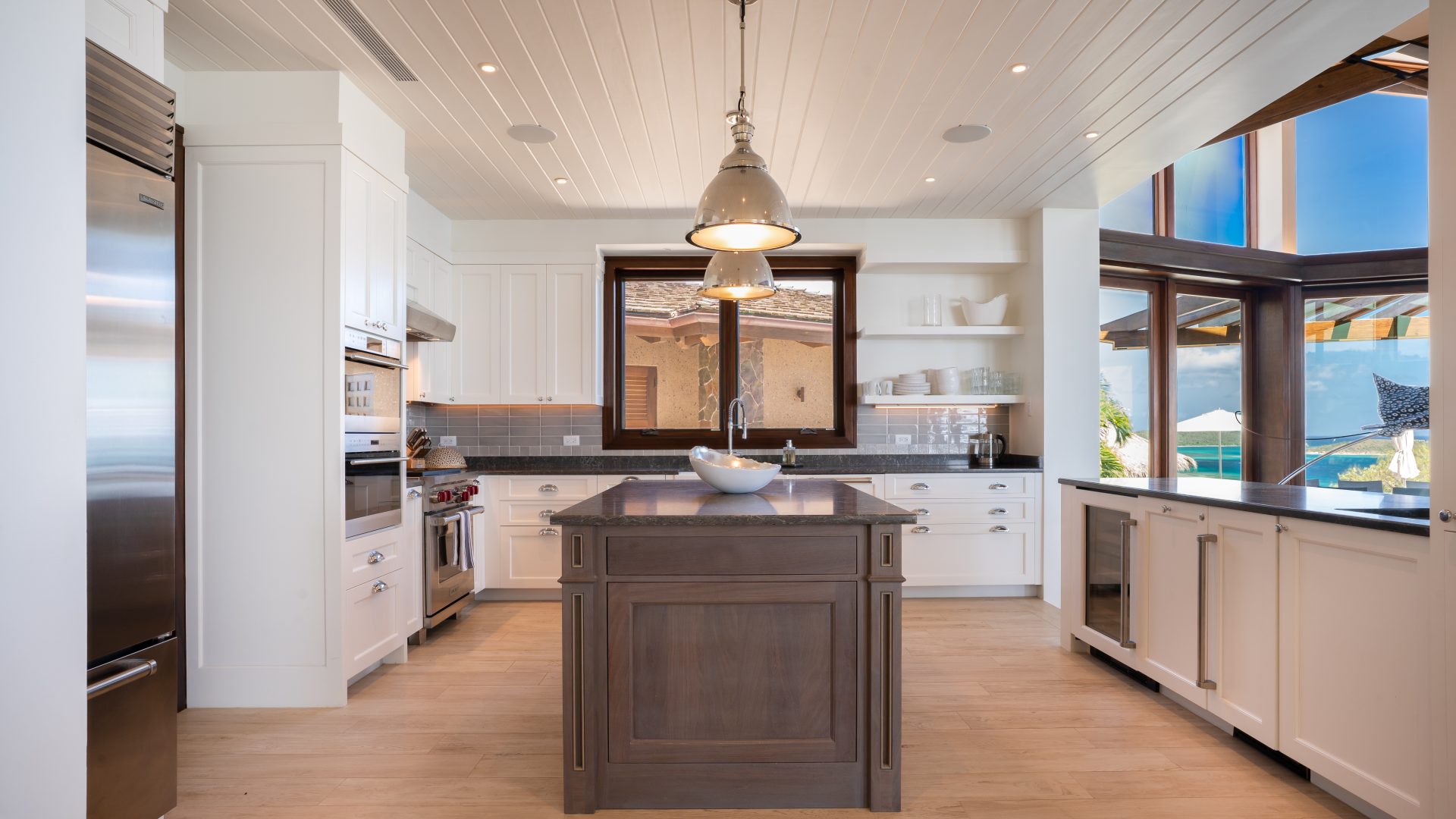 A bright, modern kitchen at Compass Rose Villa features white cabinetry, stainless steel appliances, and a dark wood island topped with a sink. Large windows offer views of an outdoor pool area. The space includes wooden flooring and ceiling, with pendant lights hanging over the island.