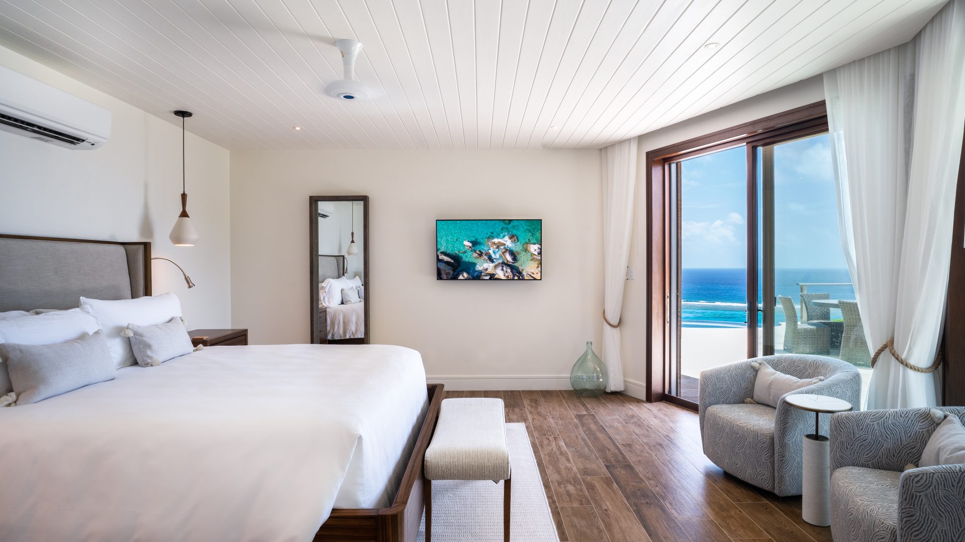 A modern, light-filled bedroom at Compass Rose Villa features a large bed with white linens, a wooden floor, and a flat-screen TV mounted on the wall. There is a full-length mirror next to a wooden nightstand. A sliding glass door opens to a balcony with ocean views. Comfortable seating is provided by two cushioned chairs.