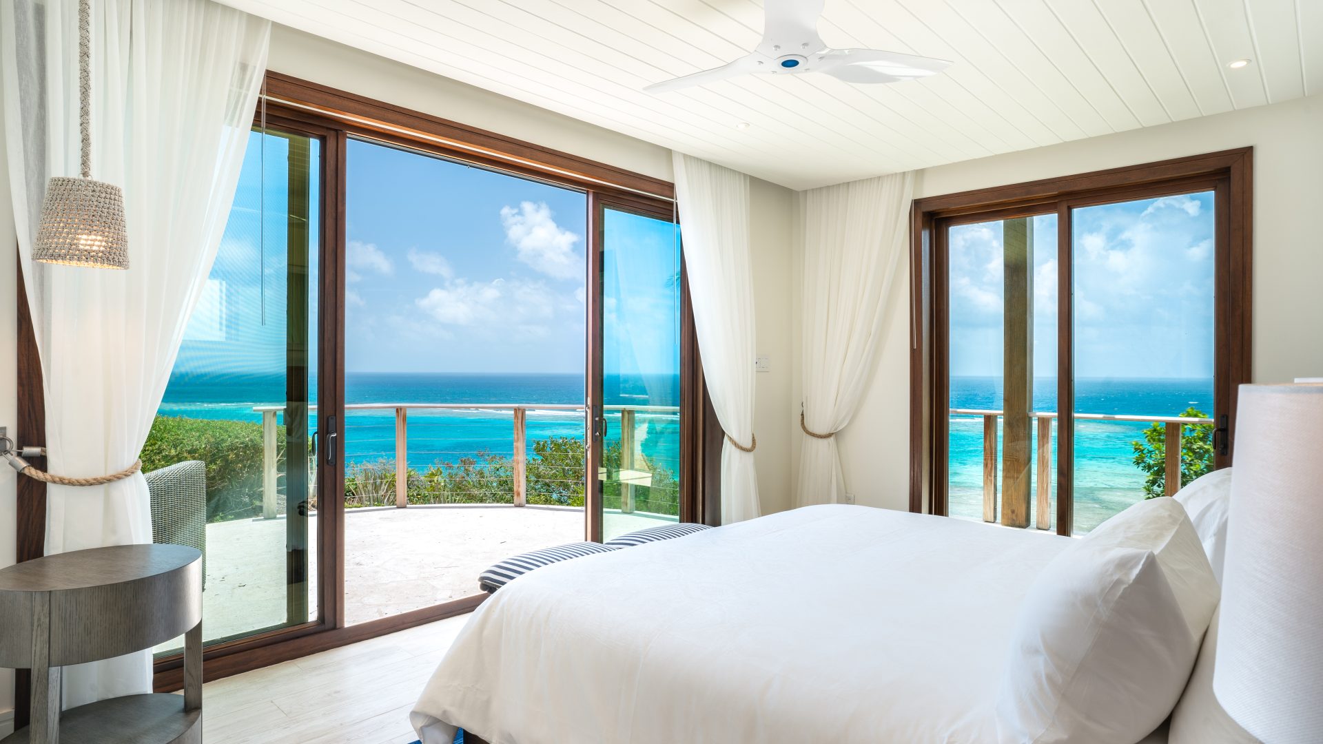 A bright, modern bedroom at Compass Rose Villa features white decor and large windows offering a stunning view of the ocean. There is a double bed, a side table with a lamp, and a ceiling fan. Sliding glass doors open to a balcony with clear blue waters and a sunny sky in the background.