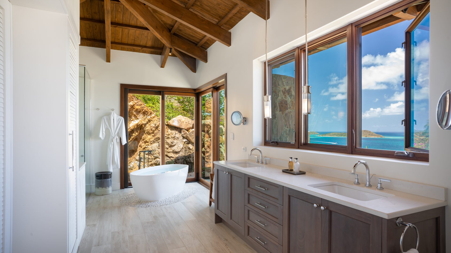 Compass Rose Villa boasts a bathroom with a freestanding bathtub near large glass doors opening to a seaside view. The interior features wooden beams, high ceilings, a double sink vanity, and a robe hanging on the wall. Outside, there's a rocky landscape and blue ocean water under a clear sky.