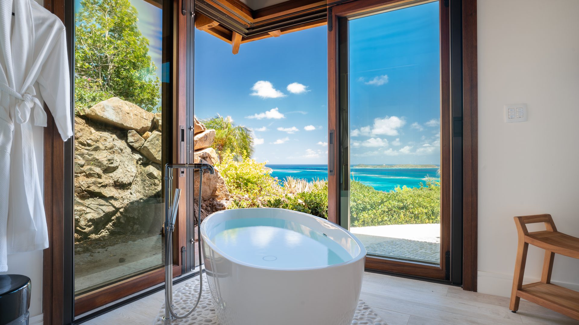 At Compass Rose Villa, a modern bathroom features a freestanding oval bathtub positioned next to large, open glass doors that offer a stunning view of a sunny, tropical ocean landscape. The room is brightly lit with natural light and exudes a serene, luxurious ambiance.