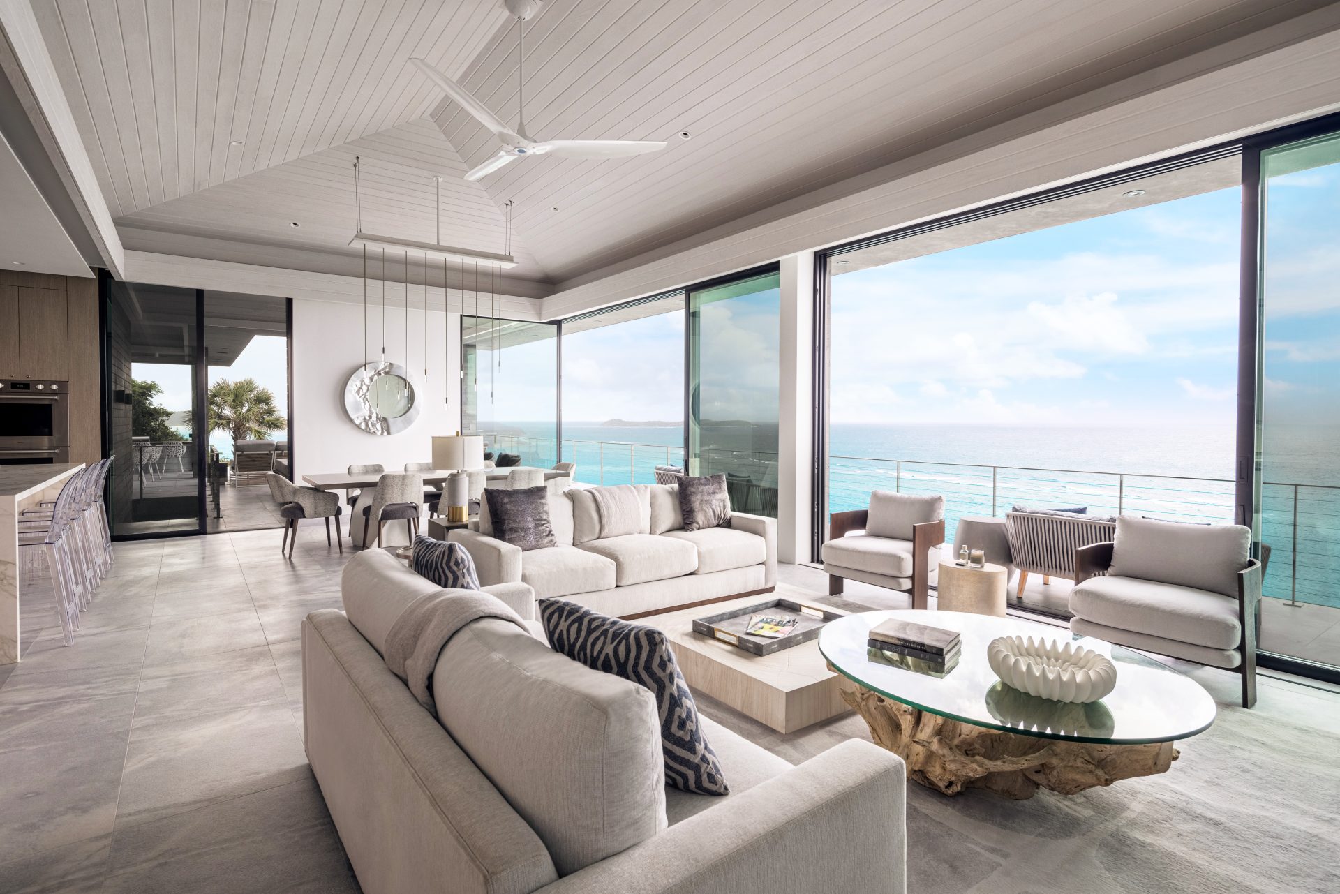 A modern living room in a luxurious villa features a high ceiling, furnished with beige sofas, armchairs, and a rustic coffee table. Floor-to-ceiling glass sliding doors open to a balcony with an ocean view. The room blends contemporary and natural decor elements seamlessly.