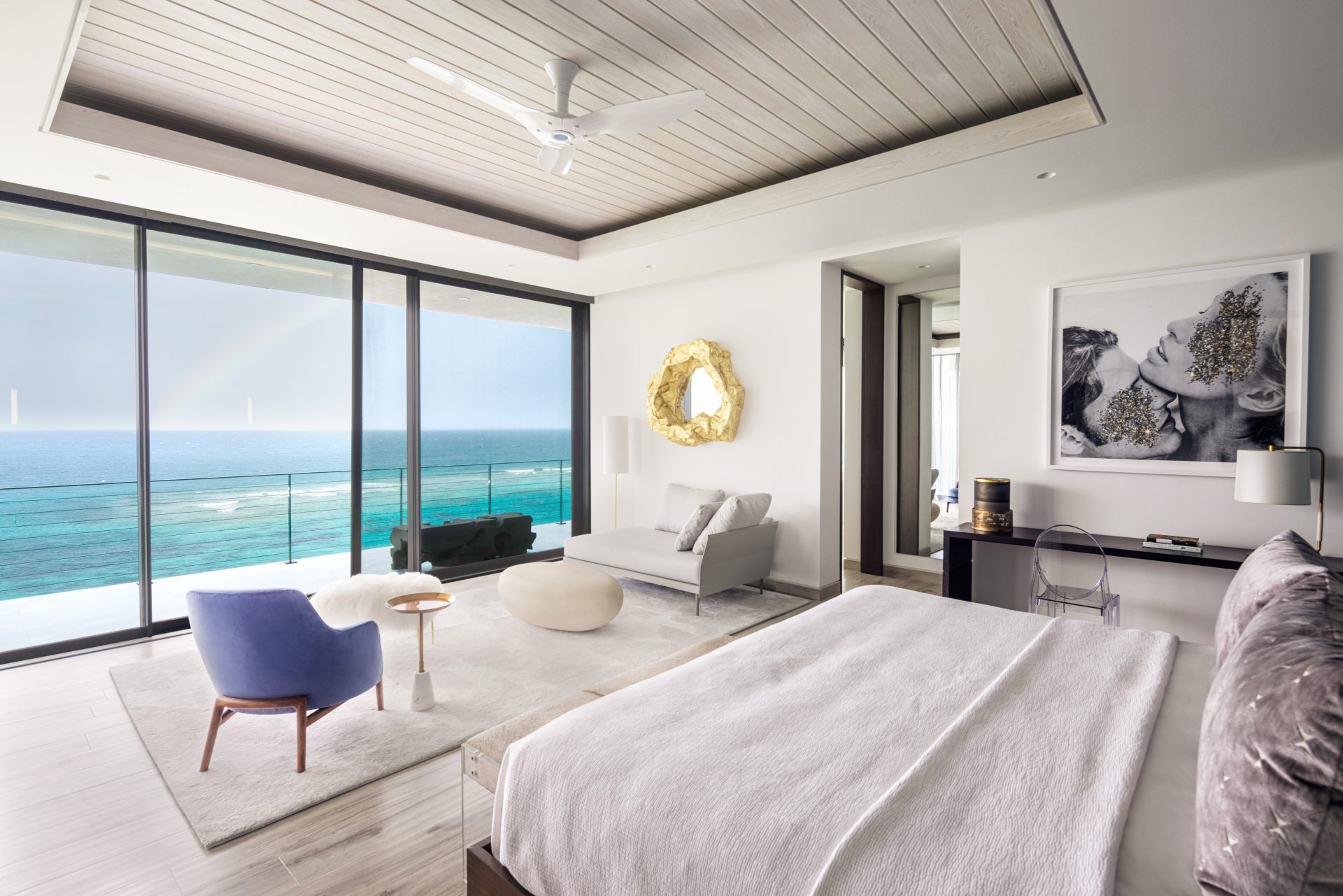 A modern bedroom in a villa features a large bed with a white coverlet, an elegant seating area with a gray sofa and a blue chair, and floor-to-ceiling windows that open to a balcony with a stunning ocean view. A golden mirror and a black-and-white portrait adorn the walls like constellations of style.