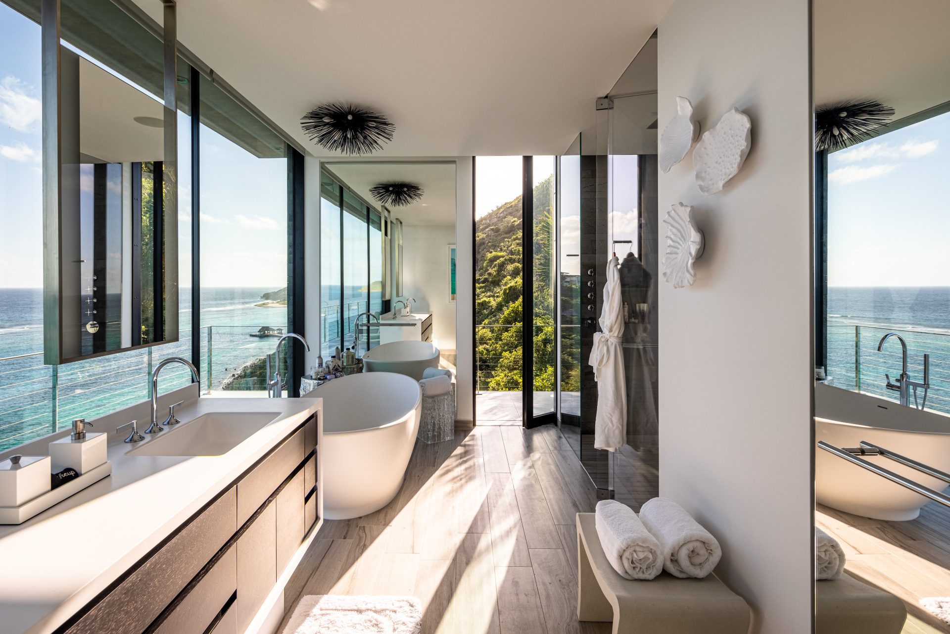 A modern bathroom in Constellations Villa boasts large windows offering a stunning ocean view. The space features a white bathtub, a double sink vanity with black cabinetry, white towels neatly rolled on a bench, and artistic wall decor. Natural light floods the room, enhancing its serene ambiance.