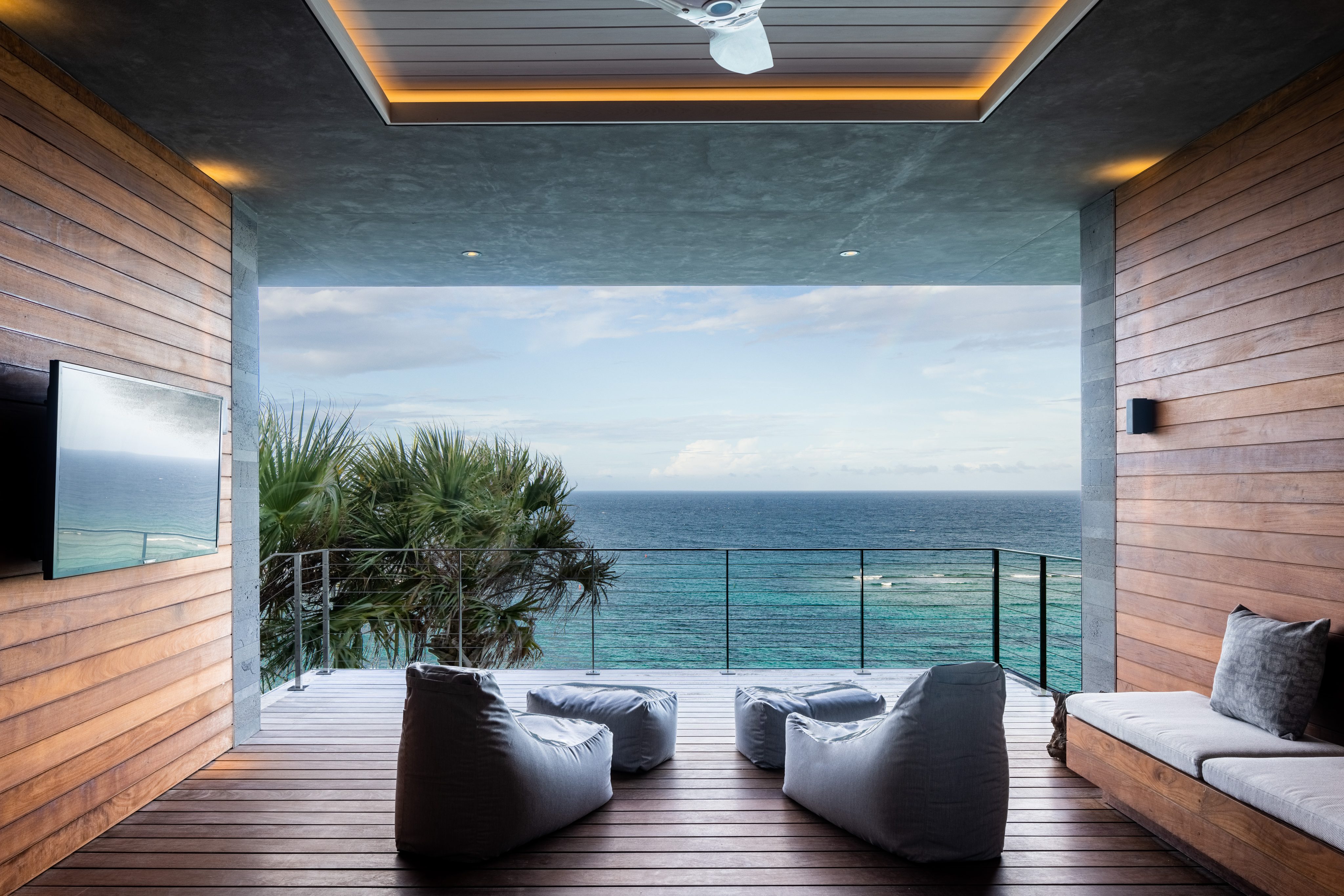 A modern, open-air room with wooden walls overlooks a breathtaking ocean view at the villa. The space features a ceiling fan, a wall-mounted TV, four bean bag chairs, and a cushioned bench. Lush greenery and the expansive sea and sky create a serene backdrop perfect for stargazing constellations.