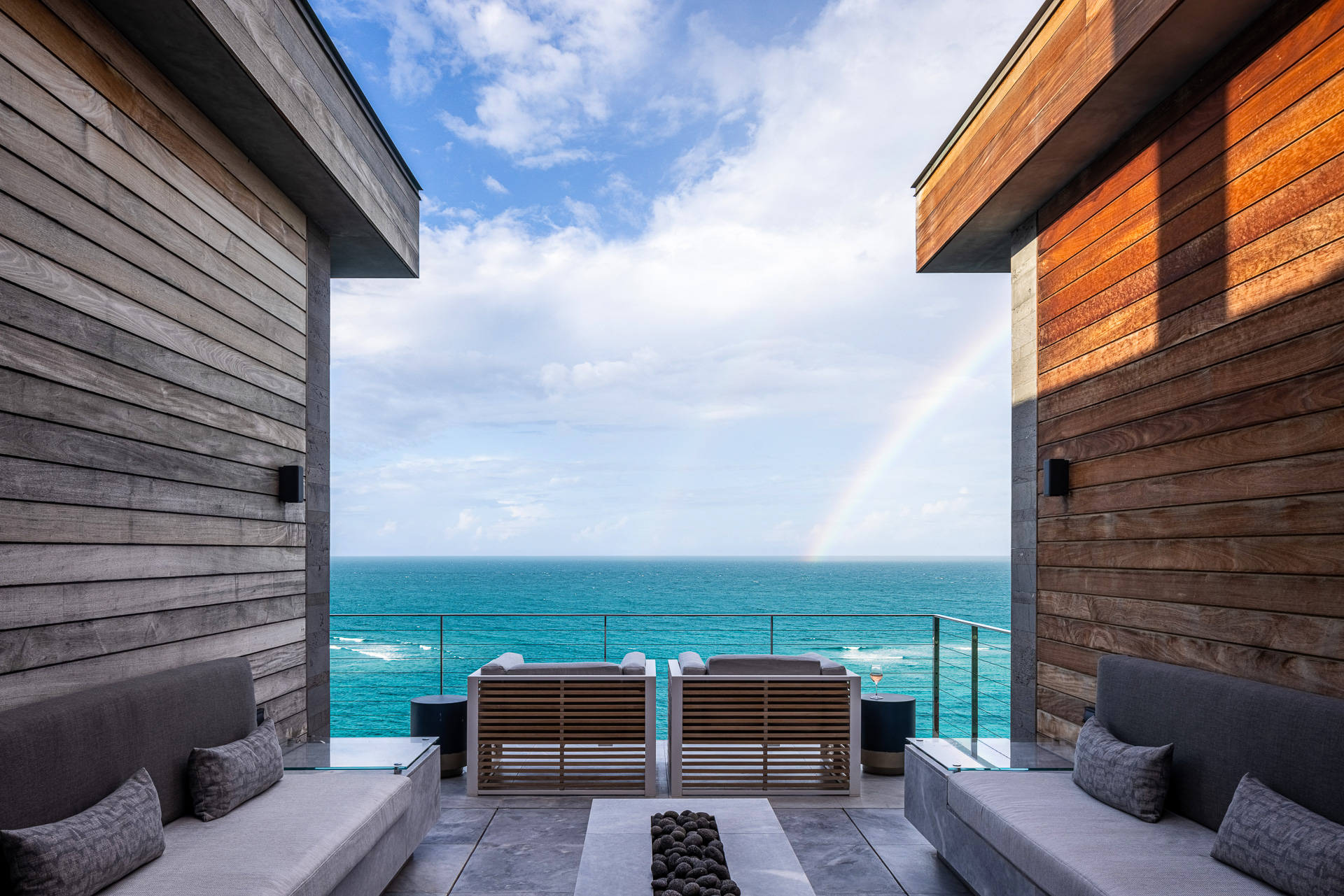 A serene ocean view is framed by modern wood-paneled walls and a cozy outdoor seating area at the villa. Two parallel wooden structures lead the eye to the horizon, where a rainbow arches over the blue sea, beneath a sky scattered with clouds and constellations starting to appear.