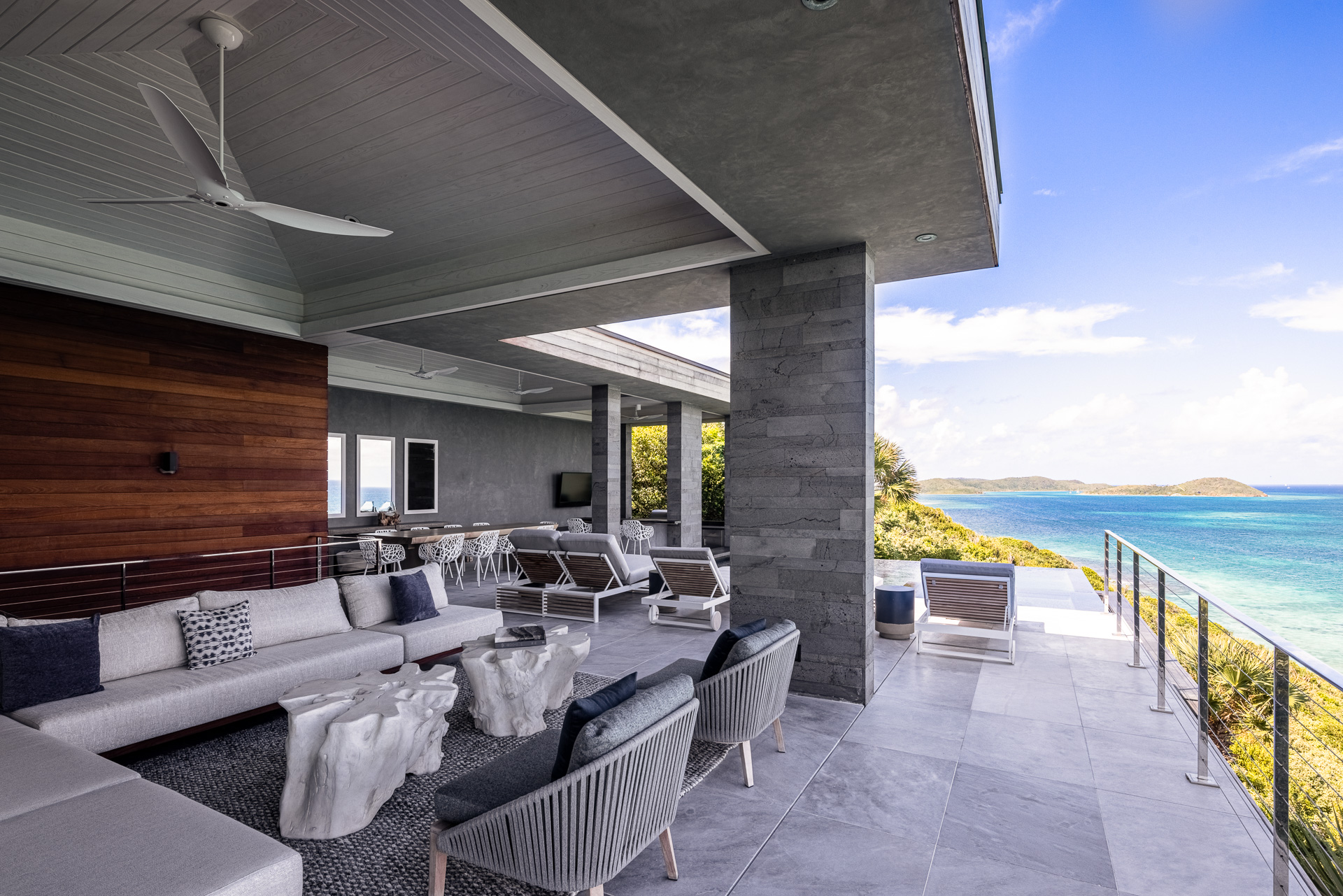 Spacious modern outdoor terrace with gray tiled flooring at the villa, stylish seating area featuring gray and white furniture, ceiling fans, and a stunning view of the ocean and distant hills under a partly cloudy sky perfect for stargazing constellations.