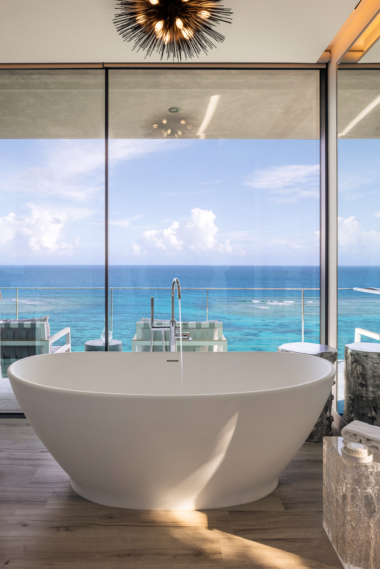 A modern bathroom in the villa features a freestanding white bathtub placed near floor-to-ceiling windows, offering a stunning view of the ocean. Outside, there's a balcony with two chairs and a table perfect for stargazing constellations. A contemporary light fixture hangs from the ceiling.