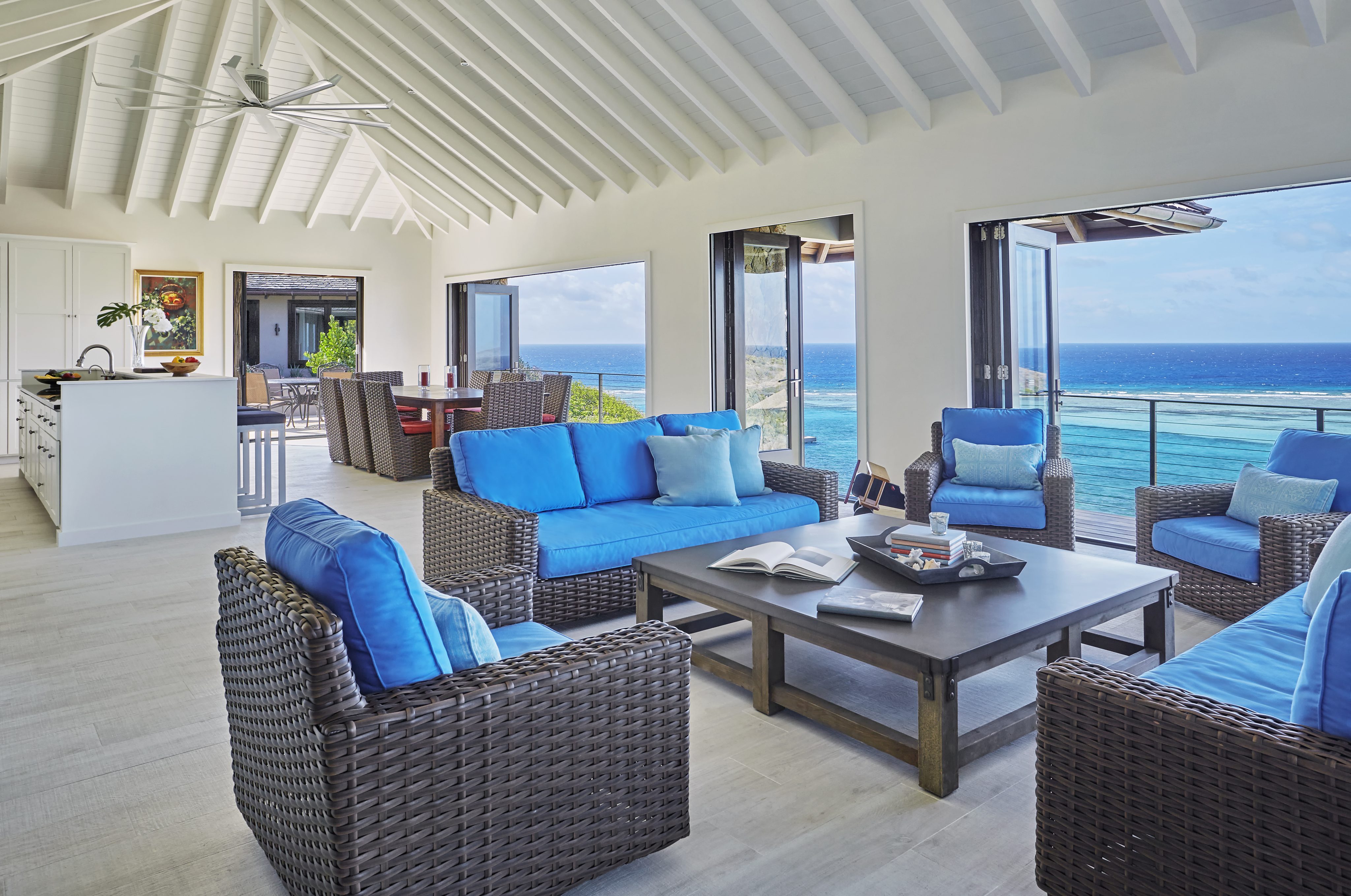 A spacious, bright living room at Cooke Lookout Villa features wicker furniture topped with blue cushions, a central coffee table with books, and a ceiling fan. Large windows provide a panoramic view of the ocean. The open layout leads to a dining area and a kitchen with a white island.