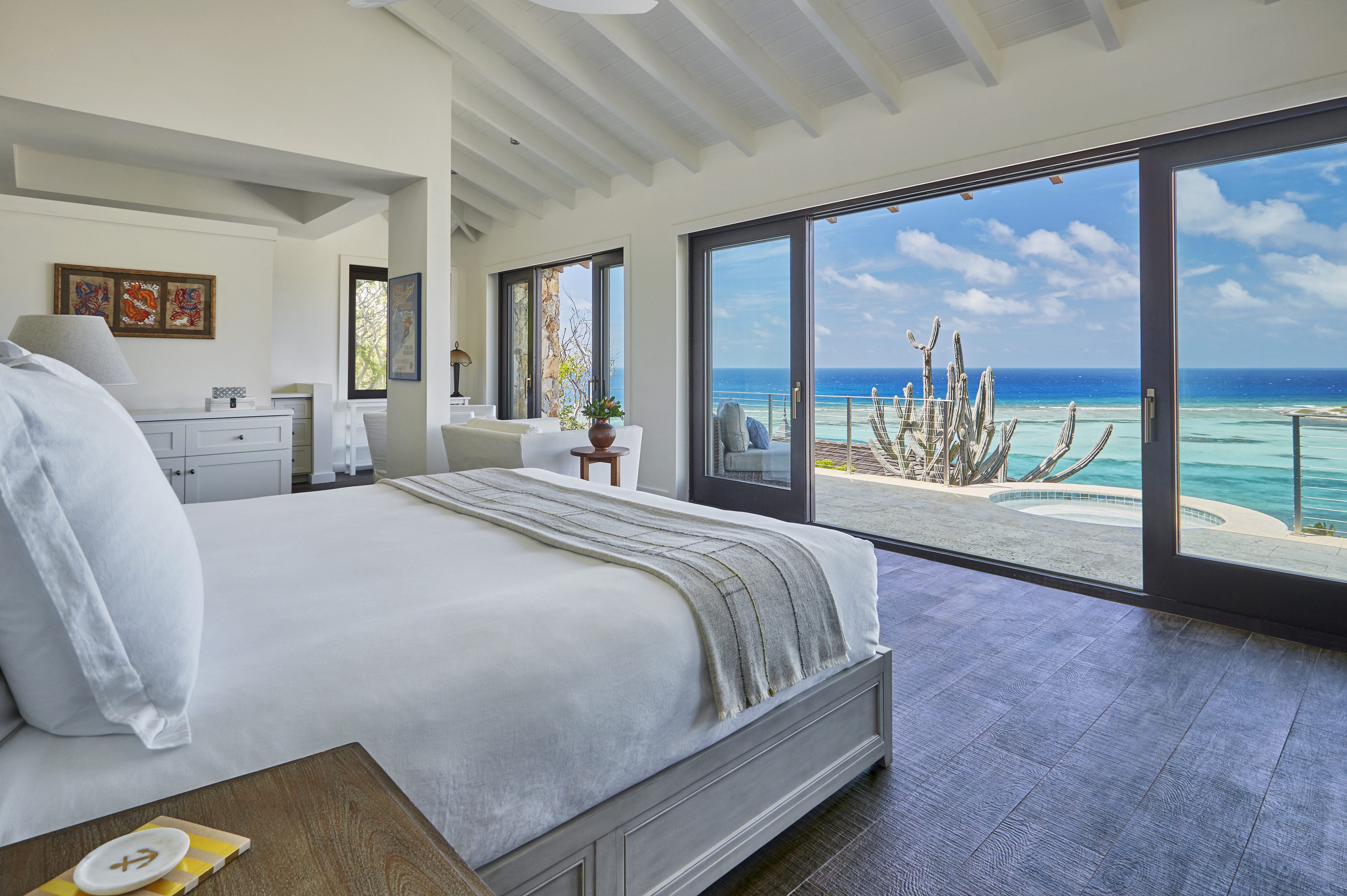 A spacious bedroom at Cooke Lookout Villa with white walls and wooden floors features a large bed with white linens and a gray throw blanket. Floor-to-ceiling windows offer a stunning ocean view, revealing a cactus sculpture, the private pool, and the clear blue sky outside.