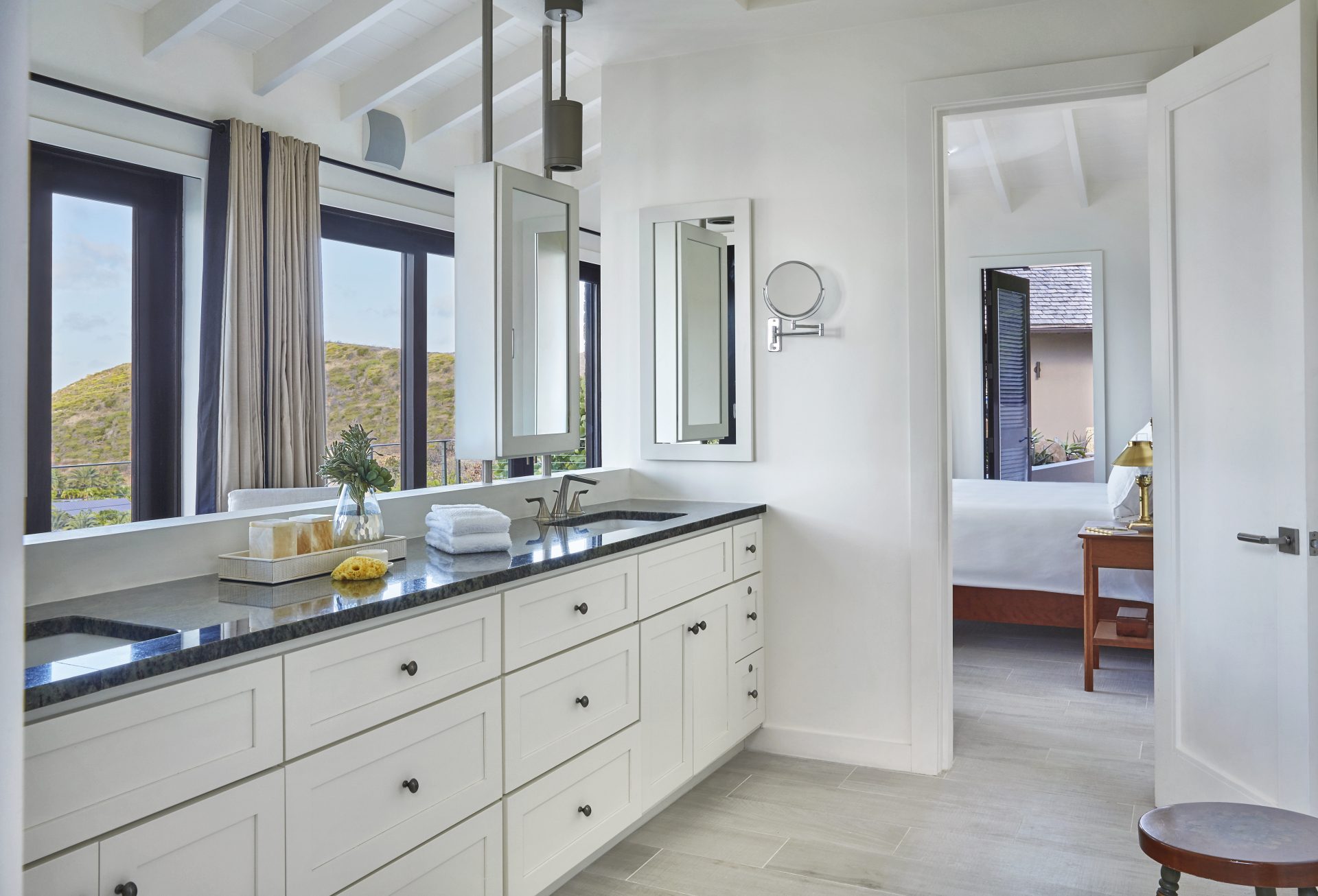 A modern, well-lit bathroom with white walls, vaulted ceilings, and large windows. The space features a long vanity with double sinks, black countertops, a makeup mirror, and various toiletries. An open doorway leads to the bedroom of this elegant villa with a bed and more windows offering a serene lookout.