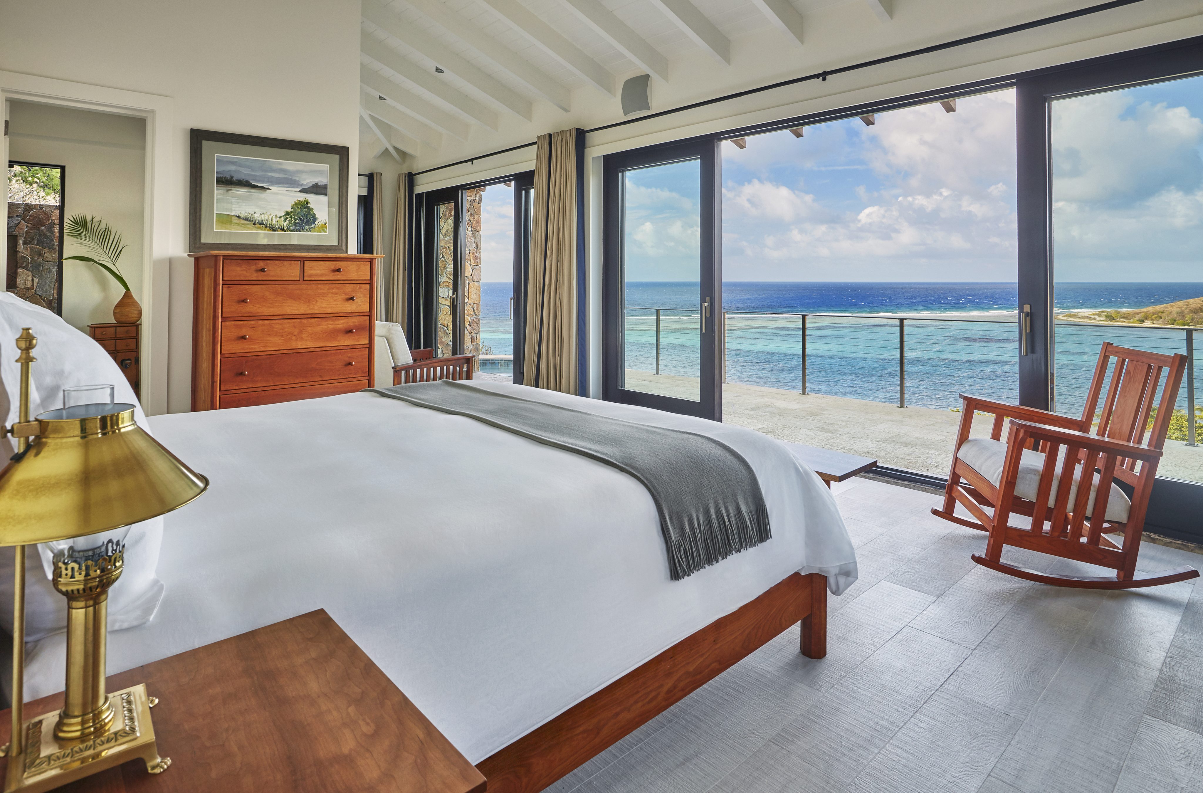 A serene bedroom at Cooke Lookout Villa features a large bed with white linens and a gray throw, a wooden dresser, and a bedside table with a lamp. Sliding glass doors open to a balcony with an ocean view. A wooden rocking chair sits near the doors, offering a peaceful spot to enjoy the scenery.