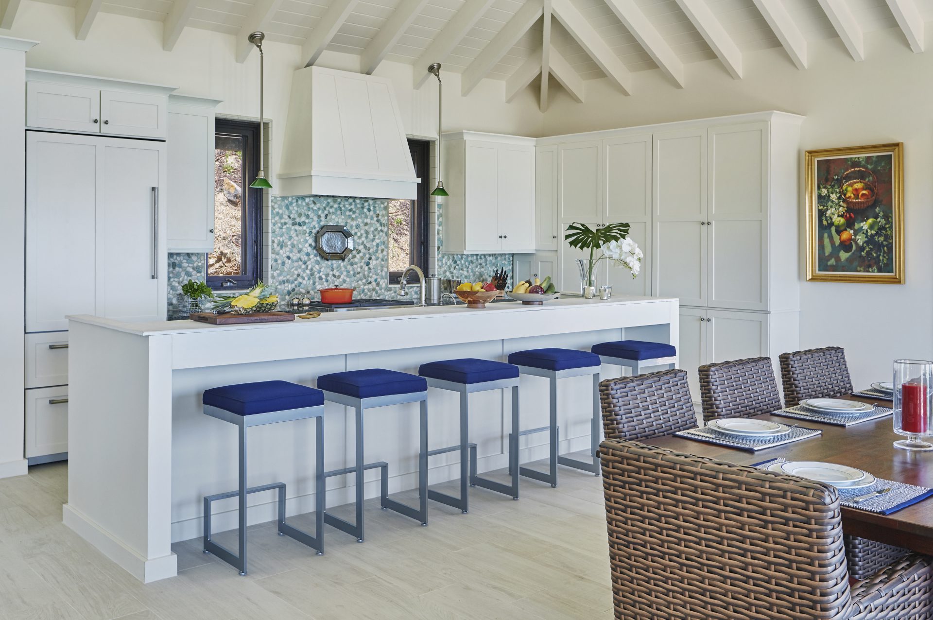 Cooke Lookout Villa features a bright kitchen with white cabinets, a large white island, and a backsplash with blue tiles. Four blue-cushioned bar stools are lined up at the island. The dining area has a wicker table and chairs. Green hanging pendant lights and a colorful painting add accents.