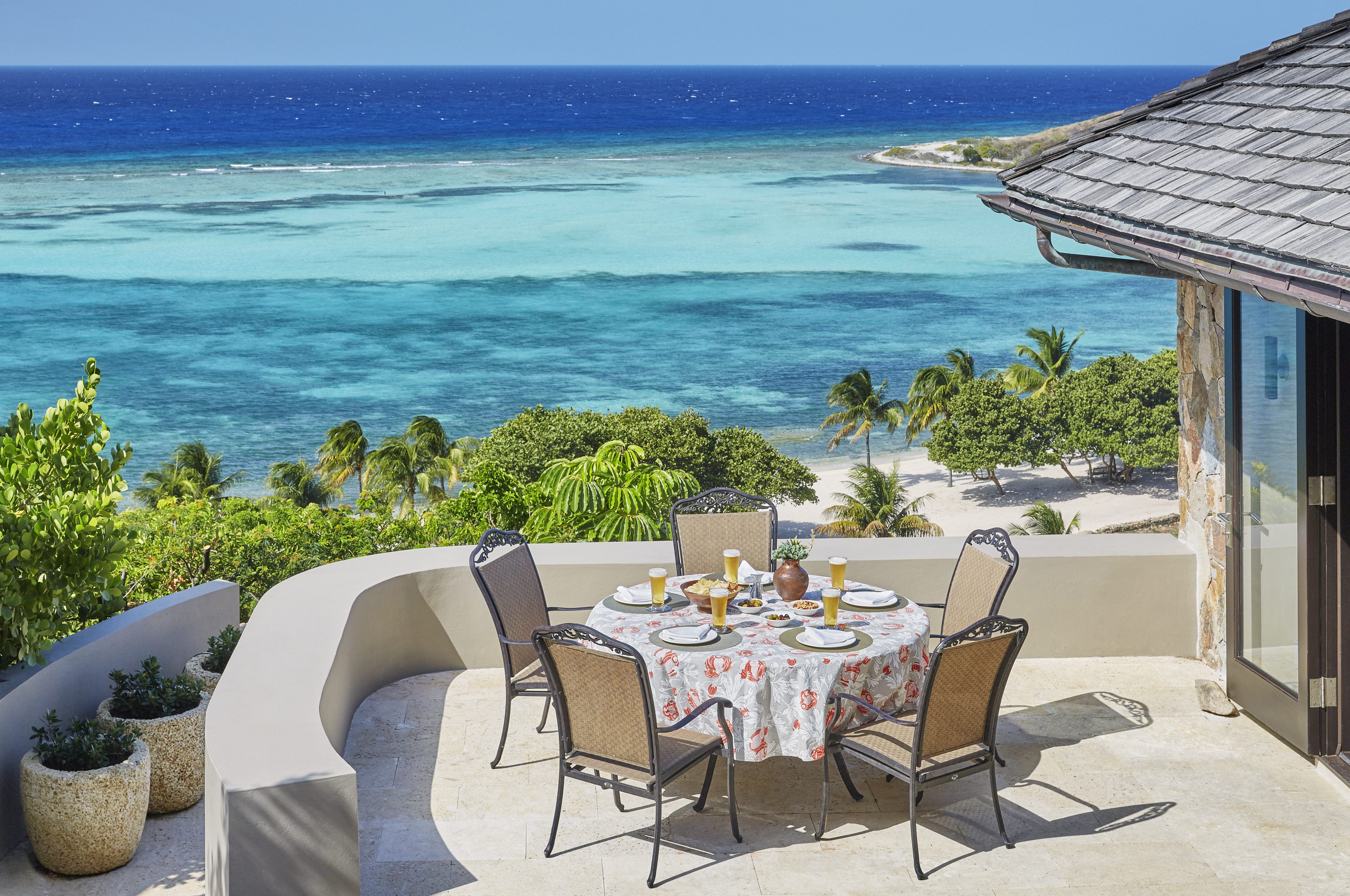 A round outdoor dining table set with a floral tablecloth, plates, cups, and cutlery graces the terrace of Cooke Lookout Villa, overlooking a tropical beach and turquoise ocean. The terrace is bordered by a light-colored wall, with lush greenery visible below near the beach.