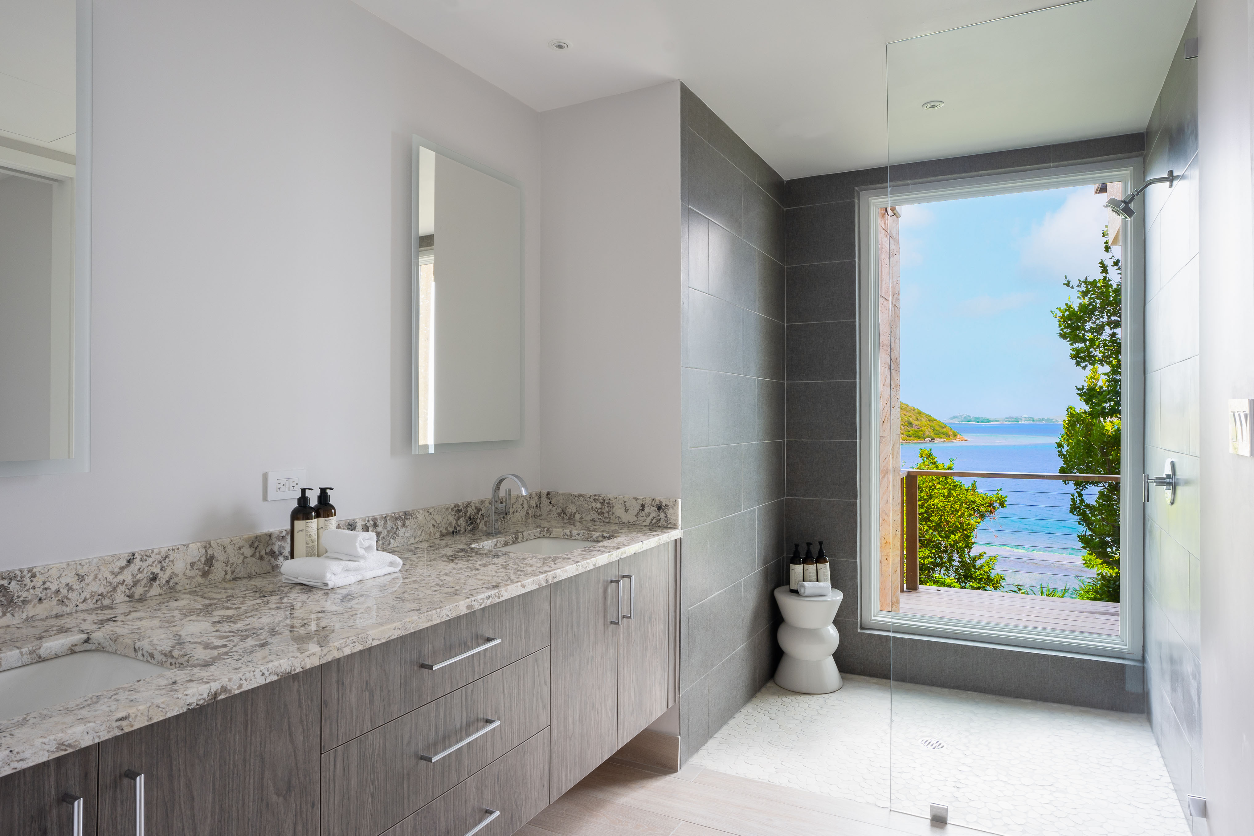 Experience the serene elegance of Deep Bay Villa’s modern bathroom, featuring a large mirror, double sink, and granite countertop. The gray tiled walls and glass-enclosed shower add to its charm, while a floor-to-ceiling window offers breathtaking views of the ocean and lush greenery.