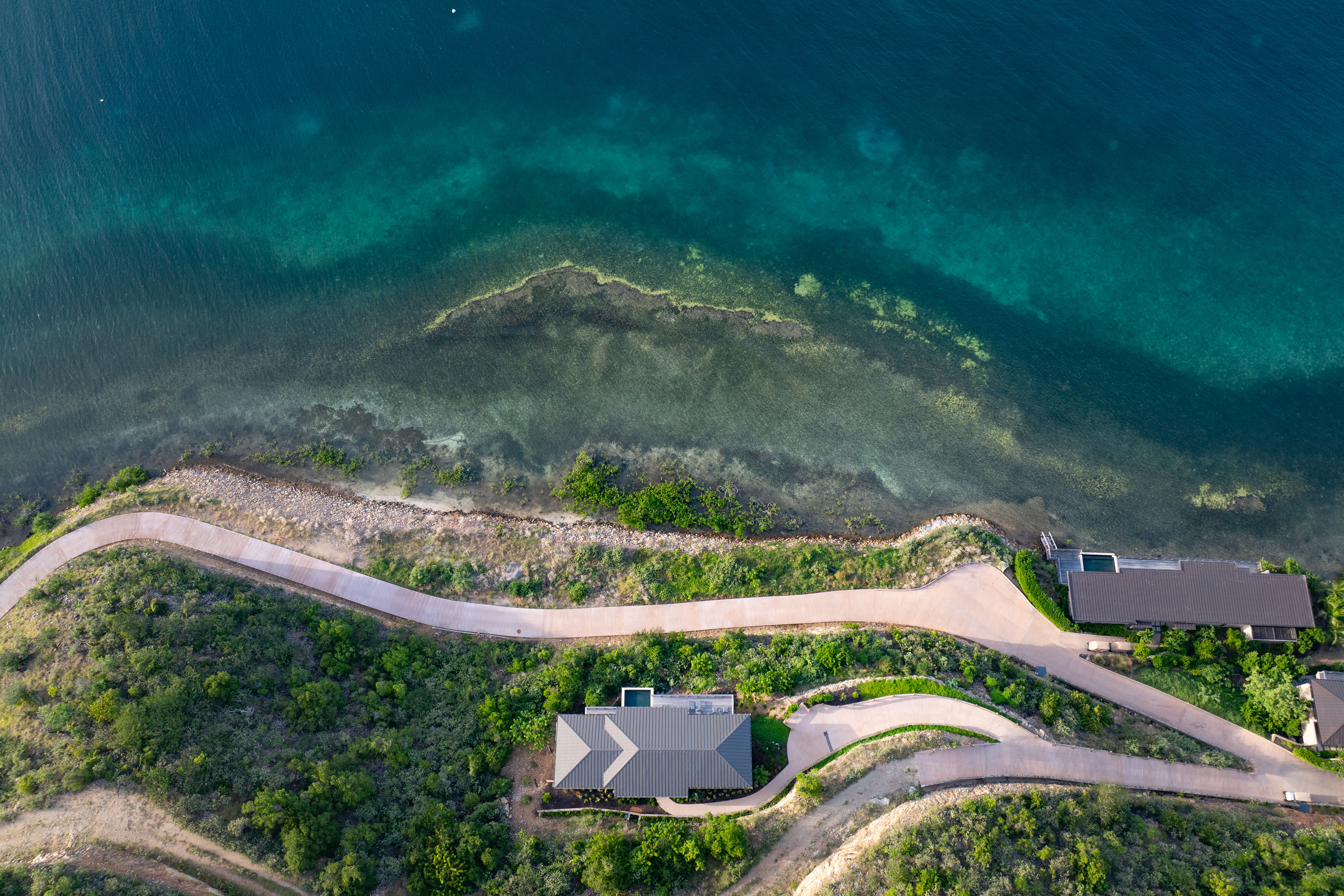 Aerial view of a coastal landscape featuring a curved road alongside a lush hillside. Deep Bay Villa is nestled by the road, with clear turquoise sea waters and rocky shoreline visible on the other side.