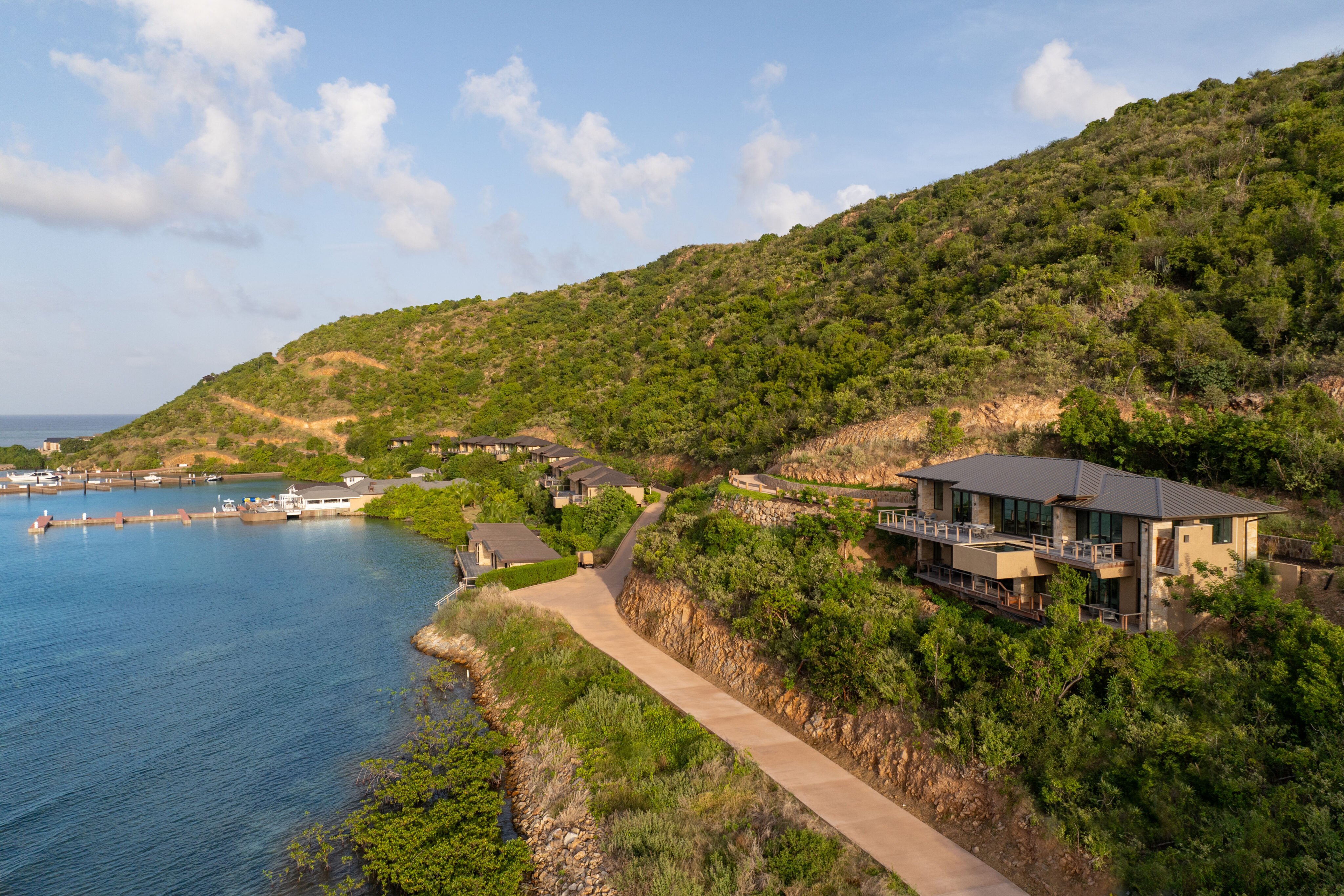 A coastal landscape featuring a winding road along a lush, green hillside with several houses, including the elegant Deep Bay Villa, perched on the slope. To the left, there is a calm body of water with boats docked at a small pier. The sky is partly cloudy.