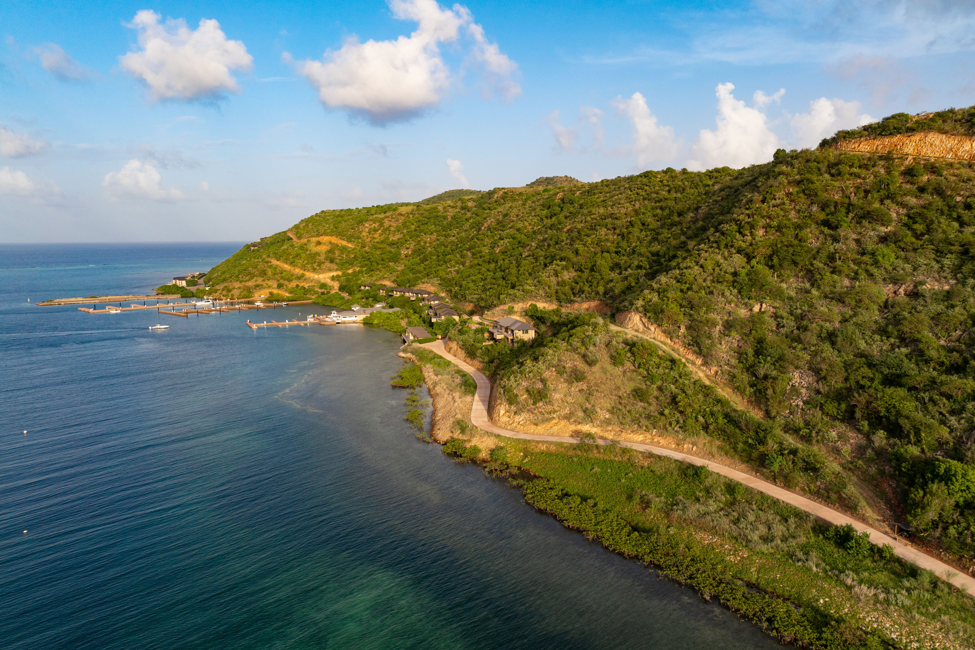 Aerial view of a coastal landscape with a winding road along lush green hills leading to Deep Bay Villa. The calm blue ocean stretches to the horizon under a clear sky, with a few clouds. A small marina with boats is visible in the distance near DB1.