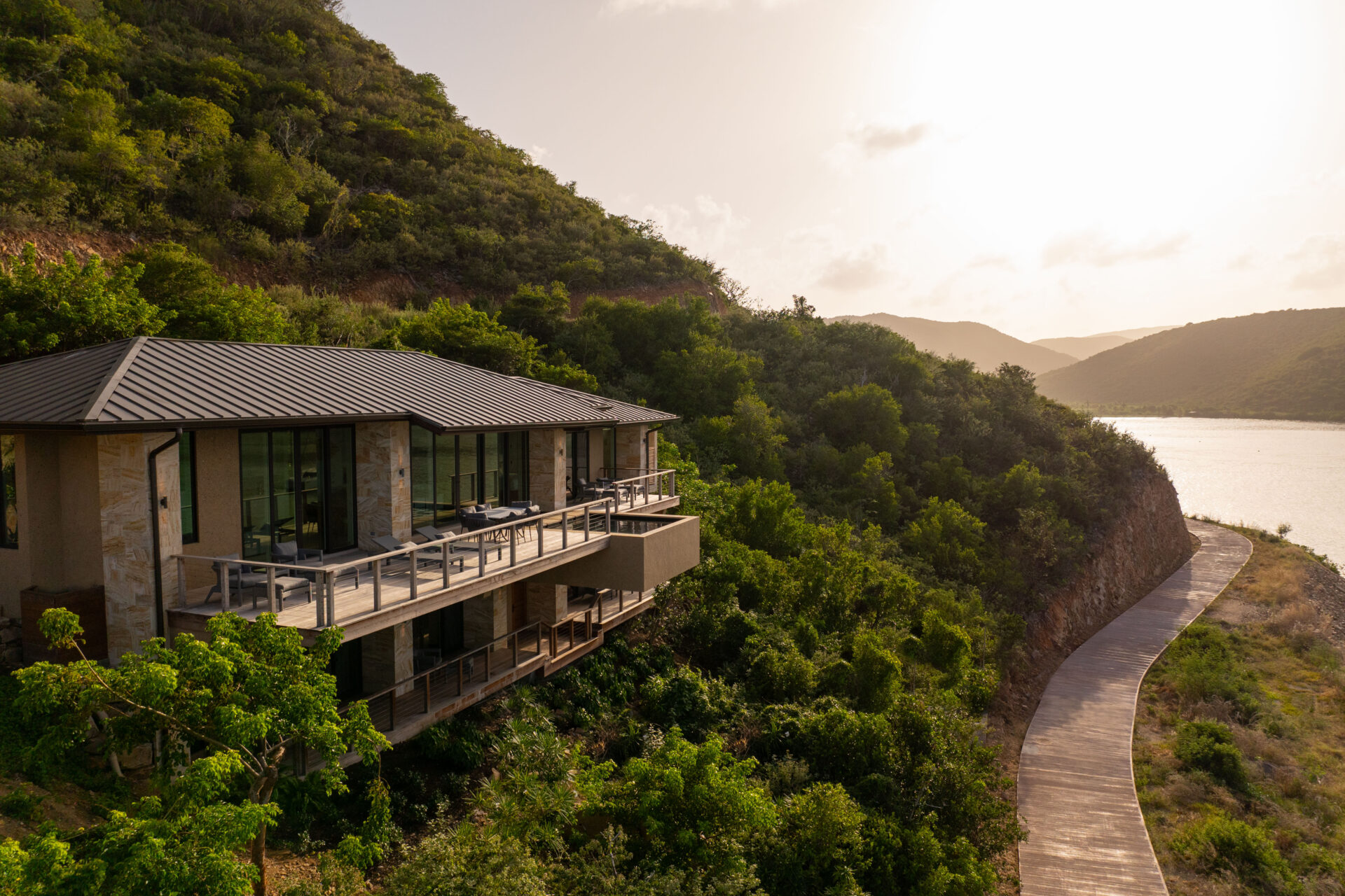 Deep Bay Villa, a modern house with large windows and a metal roof, is nestled in lush green hills. It overlooks a serene body of water at sunset. A winding wooden path leads down the hill, surrounded by dense vegetation, offering an escape for those seeking tranquility at DB1.