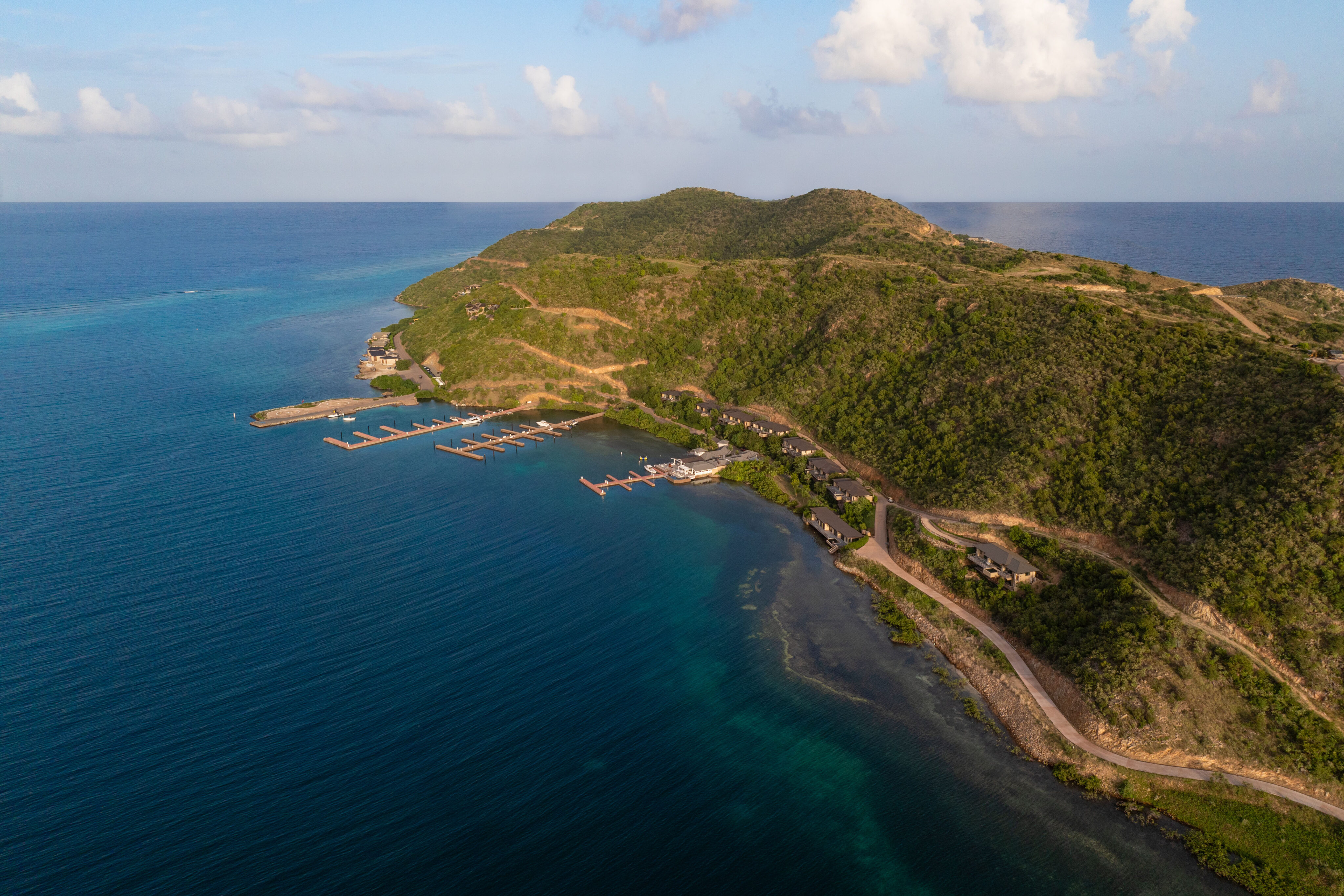 Aerial view of Deep Bay Villa on a lush, green island surrounded by clear blue ocean. The coastline, home to DB1, is lined with several docks extending into the water. A winding road leads through the hilly landscape under a partly cloudy sky.