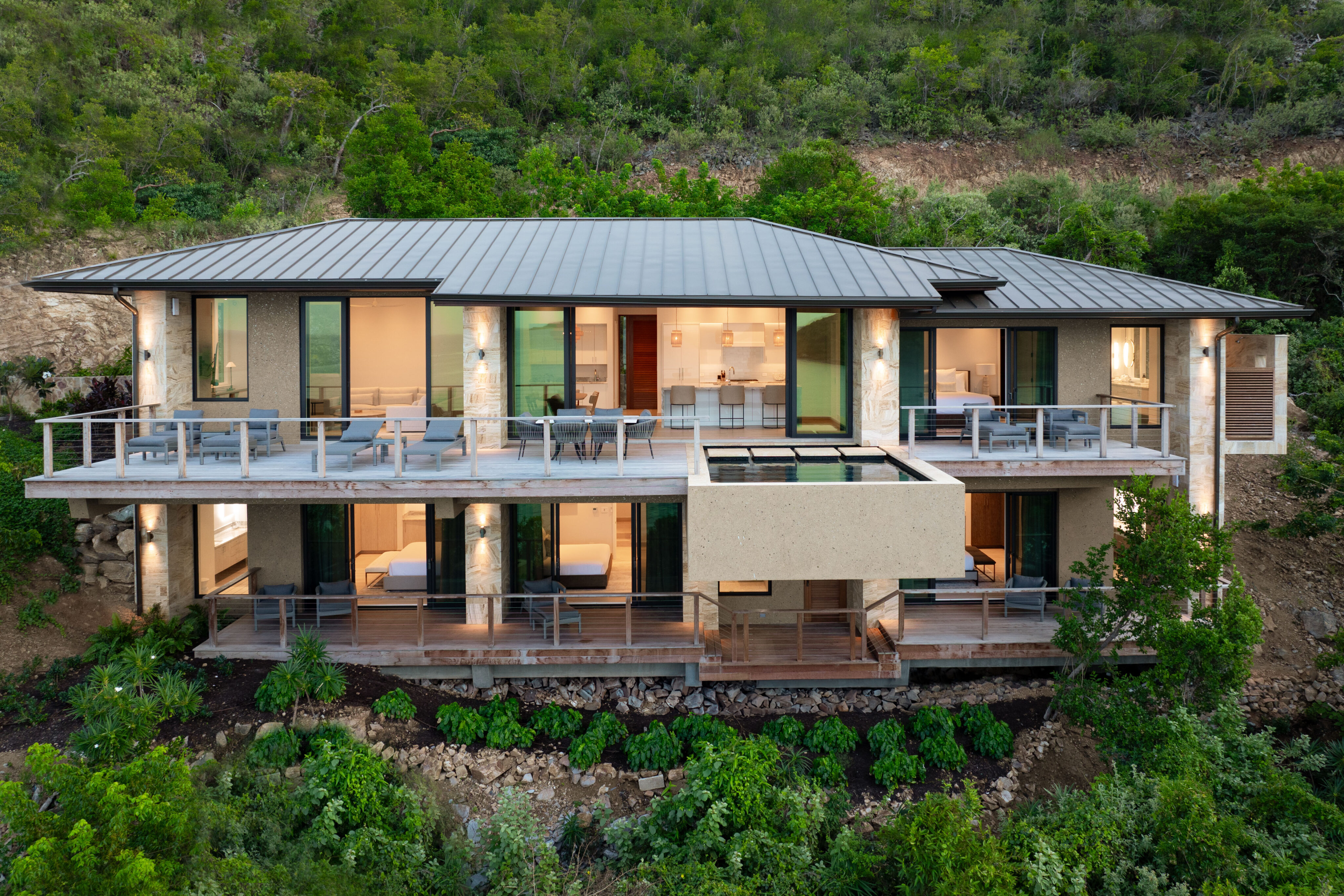 Nestled in lush greenery on a hillside, Deep Bay Villa is a modern two-story house with large glass windows and spacious balconies. The sleek metal roof and contemporary design of this DB1 structure blend seamlessly with the surrounding trees and natural landscape.