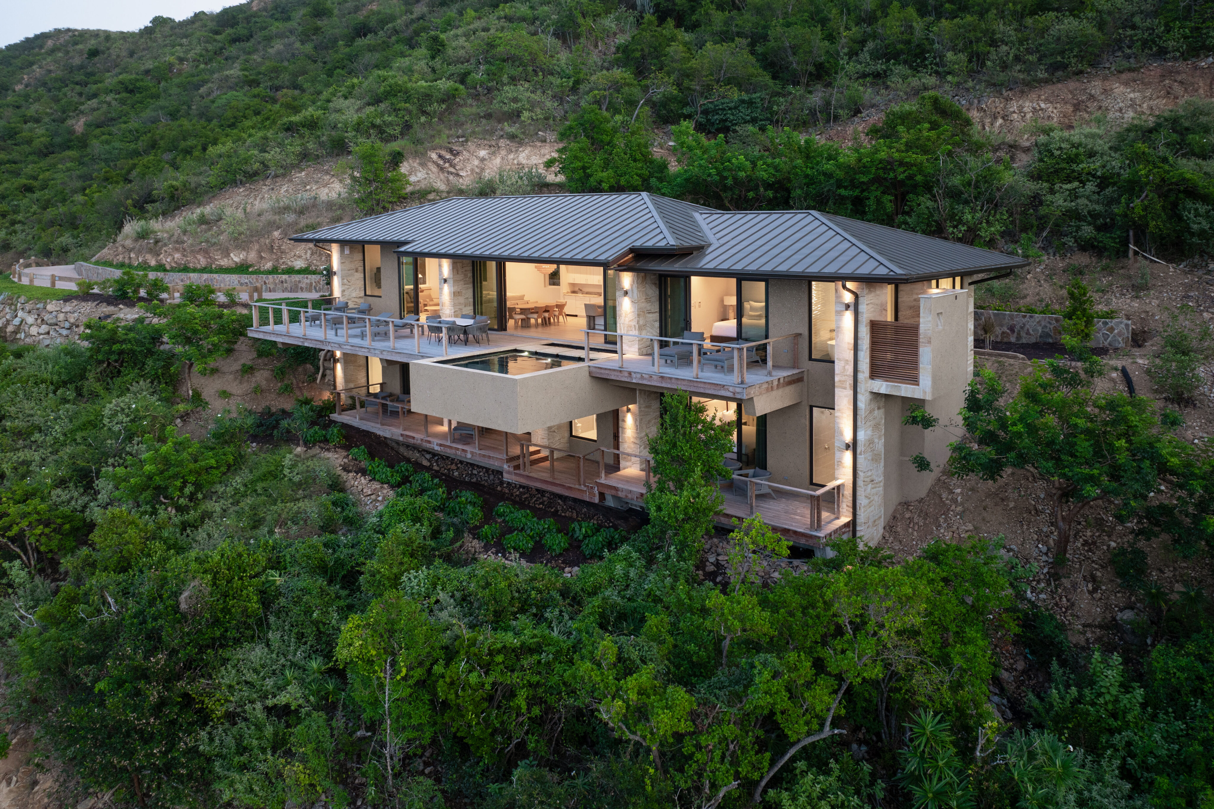 Deep Bay Villa, a modern two-story house with large windows and balconies, is nestled on a hillside surrounded by lush greenery. The architecture features clean lines and a flat roof, seamlessly blending with the natural landscape, epitomizing elegance in harmony with its surroundings.