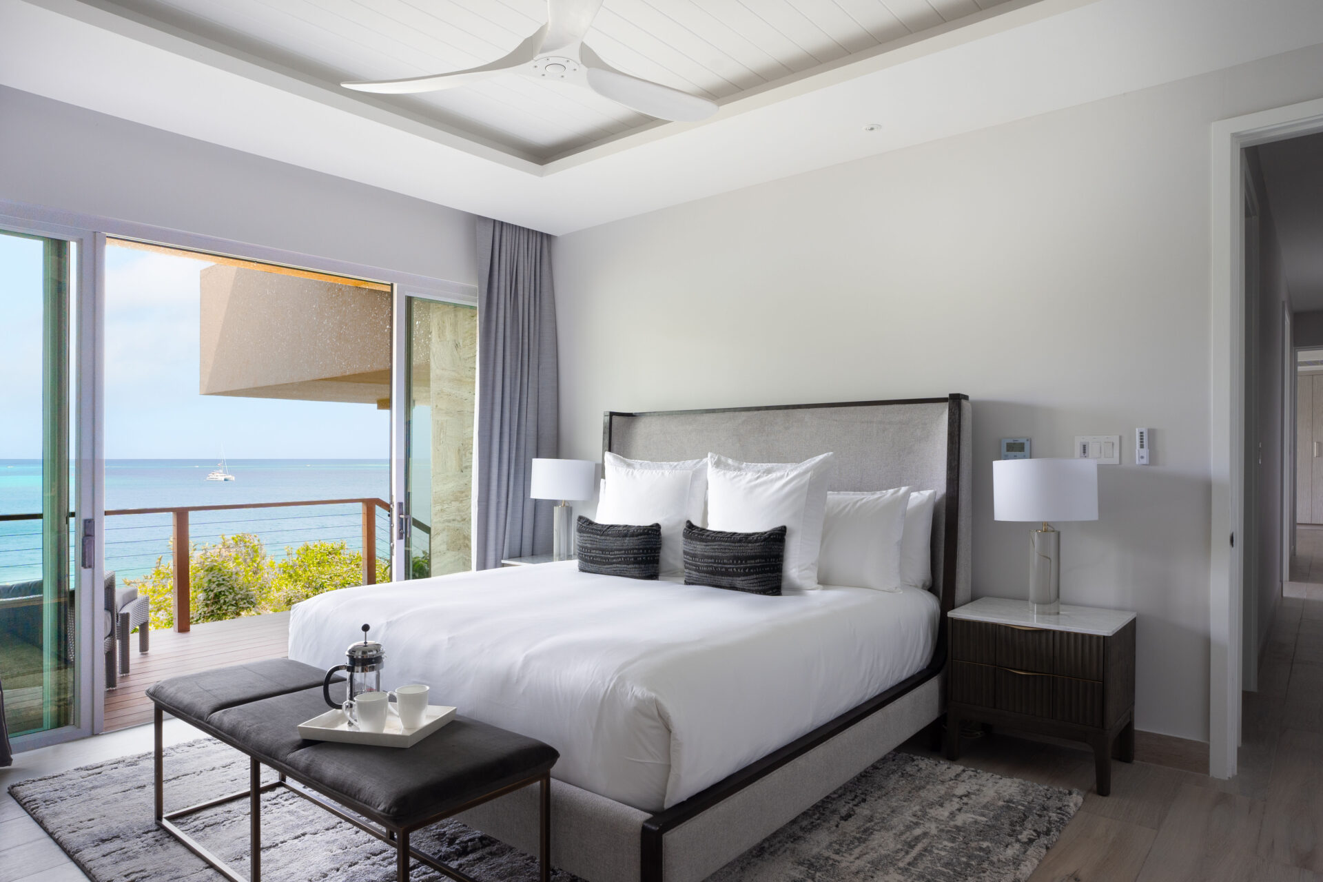 A modern bedroom in Deep Bay Villa boasts a king-sized bed and plush pillows with a balcony offering ocean views. A ceiling fan gently stirs the air, while a coffee tray on the bench invites relaxation. The large sliding glass door bathes the room in natural light, enhancing its serene ambiance.