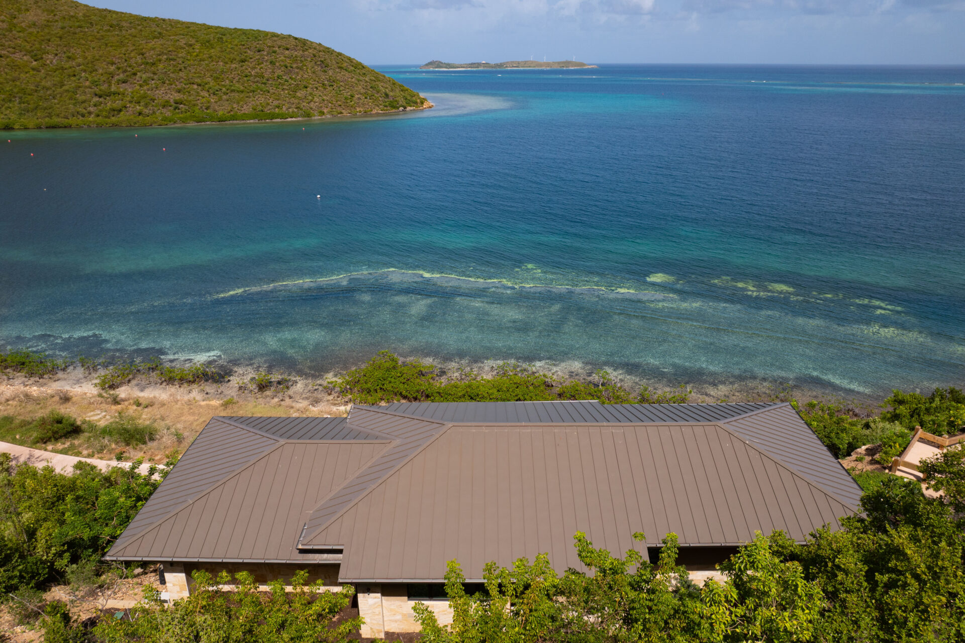 Aerial view of Deep Bay Villa with a brown roof surrounded by lush greenery. This villa overlooks a clear blue sea with small waves, and there are green hills and islands in the background under a partly cloudy sky.