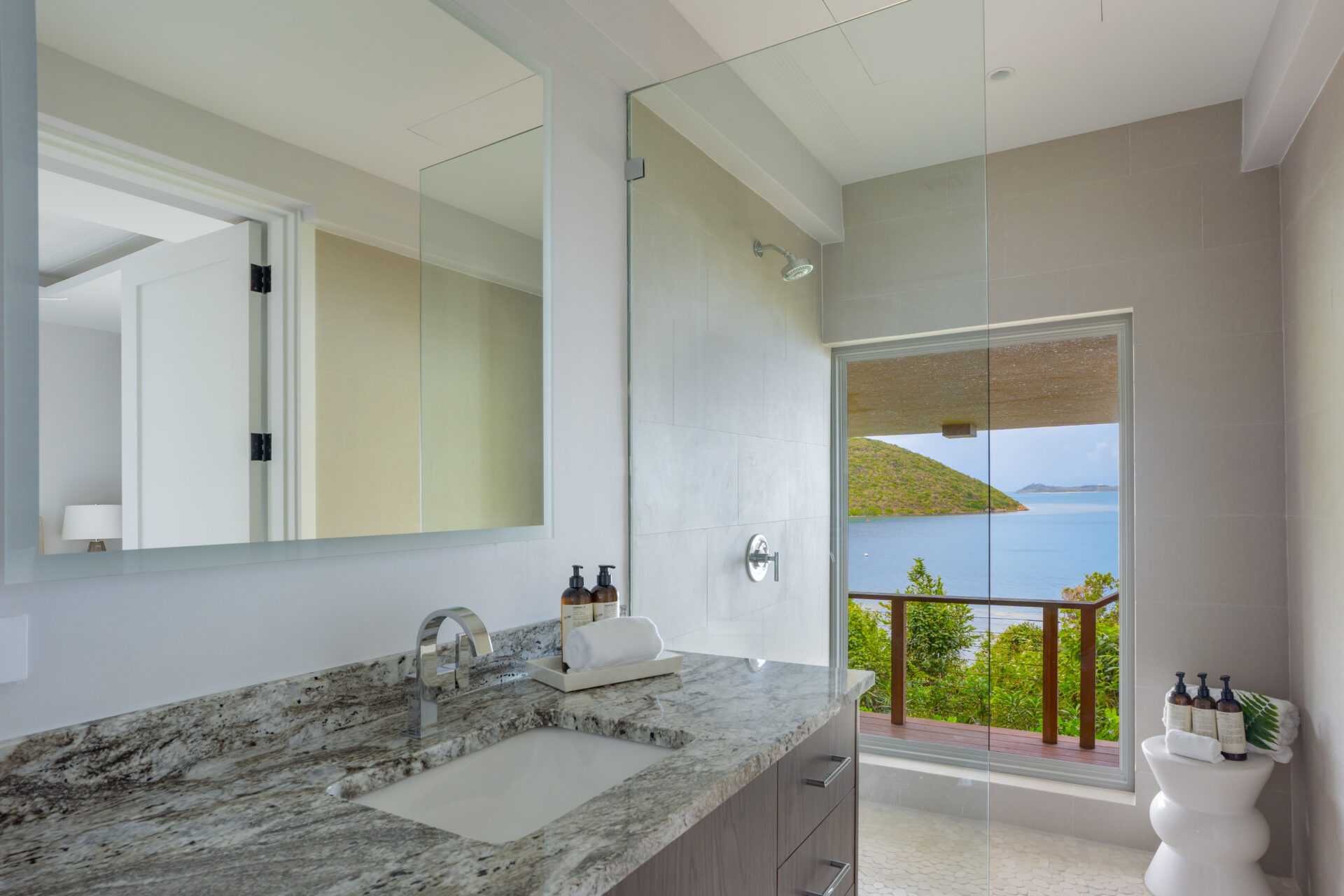 Modern bathroom in Villa DB1 with a large countertop, stainless steel faucet, and mirror. The glass shower door and window offer a serene view of Deep Bay's ocean and greenery, enhancing the tranquil atmosphere.