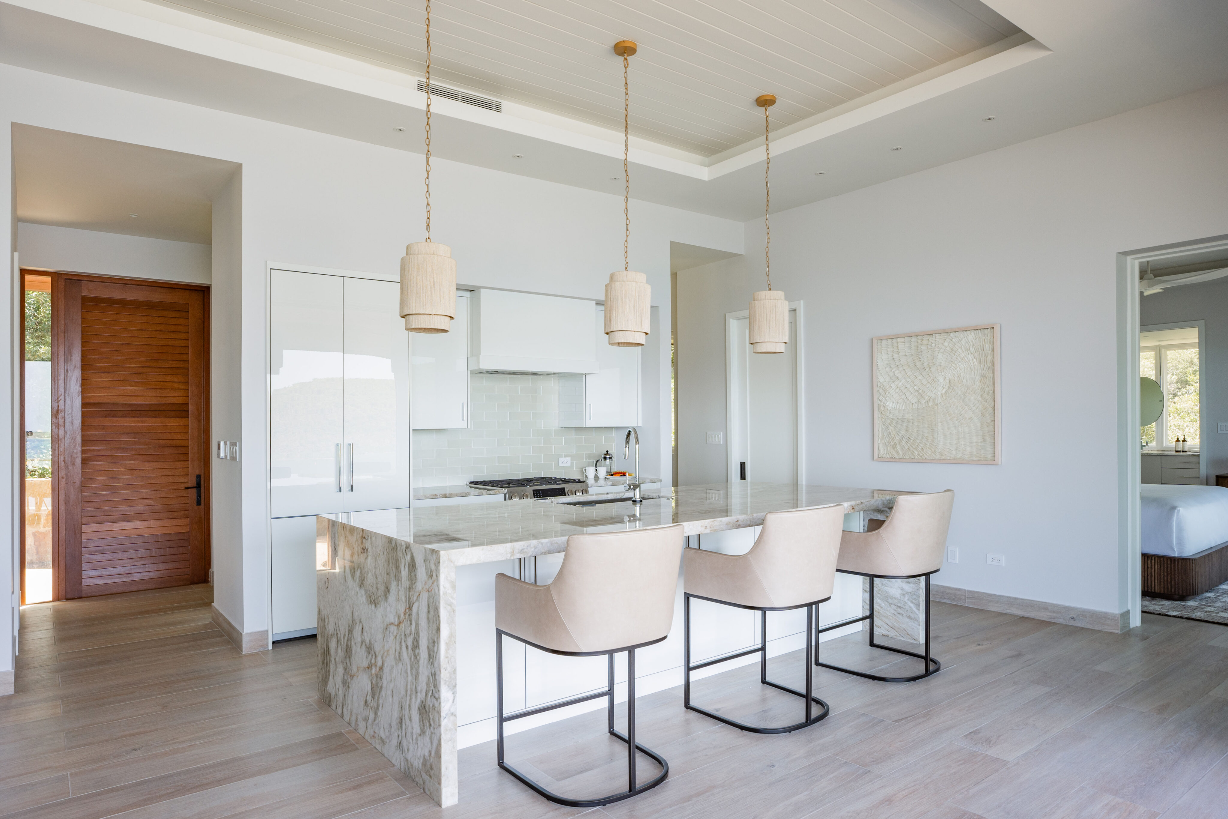 The modern kitchen in Villa DB1 features a large marble island, four beige bar stools, and pendant lights. This bright space offers white cabinets, wood flooring, and a wooden door. A textured artwork adorns the wall, while an open door reveals a glimpse into the bedroom.