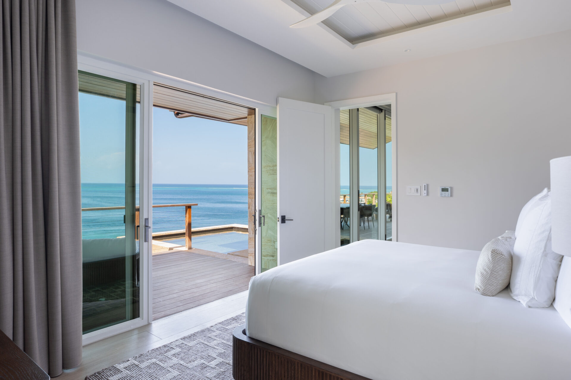 A modern bedroom in Villa DB1 features a large bed and glass sliding doors that open to a balcony with a wooden railing, offering a stunning ocean view. The room at Deep Bay Villa boasts neutral decor and an abundance of natural light, creating a serene retreat.